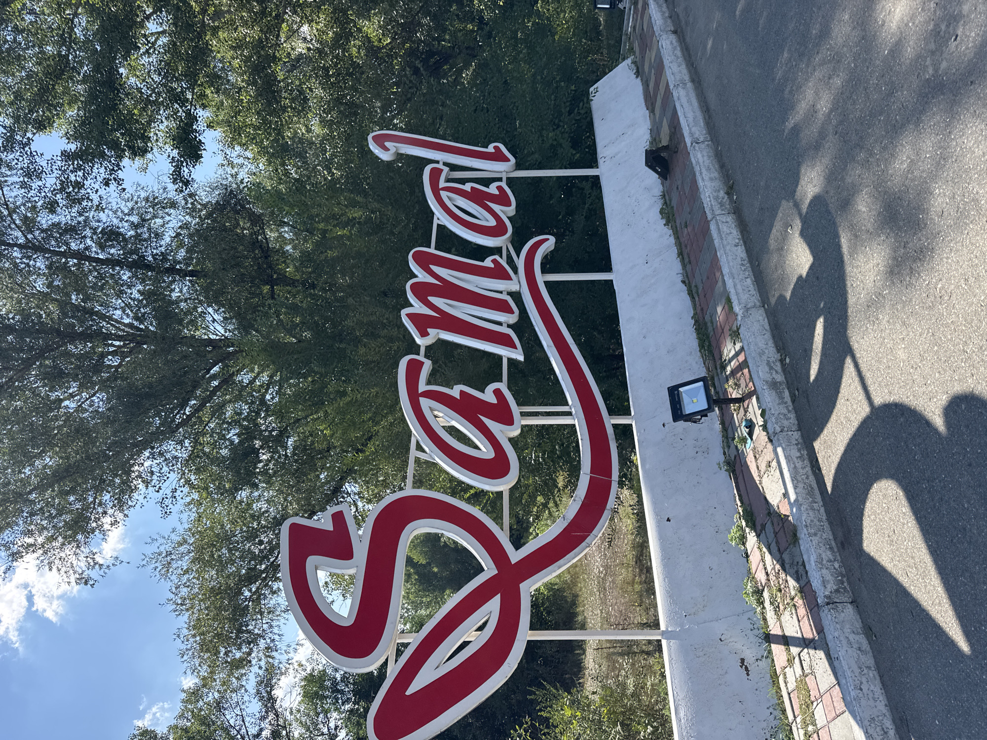 The image shows a large, red and white sign that spells out "Samal." The sign is situated outdoors, with lush green trees and a blue sky visible in the background. A white concrete barrier runs along the bottom of the sign, with a paved walkway and an asphalt road in front. A solar-powered light is mounted on the barrier. The sign appears to be at an entrance or landmark in the city of Oskemen, Kazakhstan.