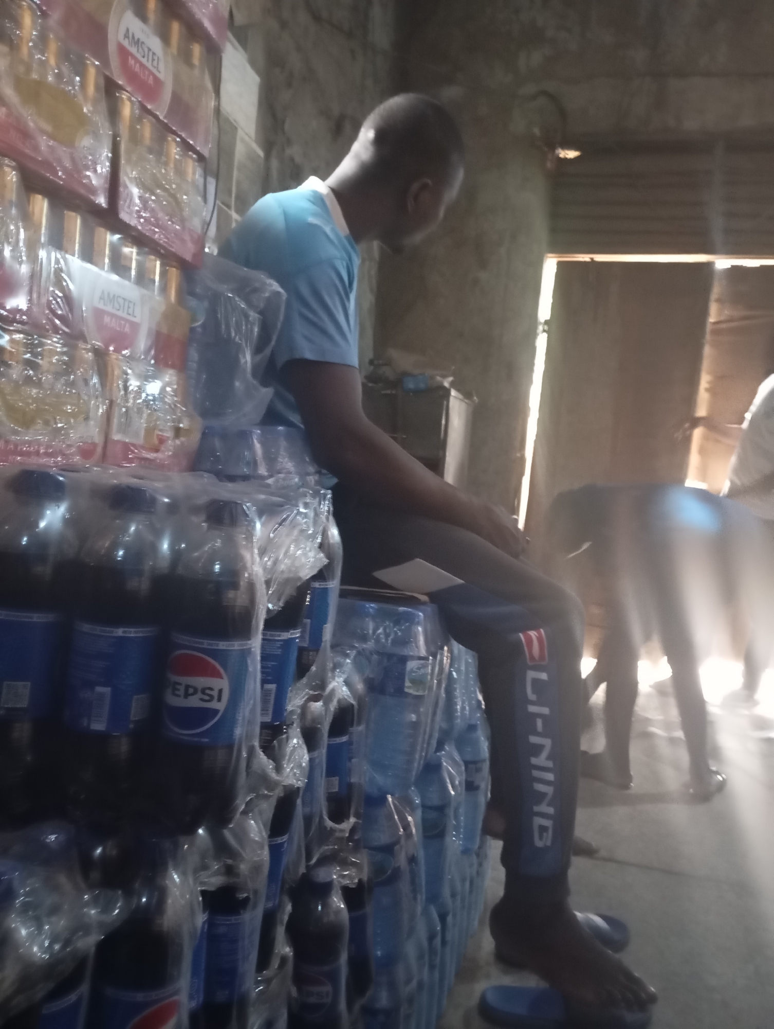 In Shendam, Nigeria, a person wearing a blue t-shirt and black pants with white lettering is seated next to stacks of bottled drinks, including Pepsi. The person is looking to the right. In the background, another person is partially visible, and there is a doorway with light coming through. The environment appears to be an indoor storage area or shop with rough-textured walls and ceiling.