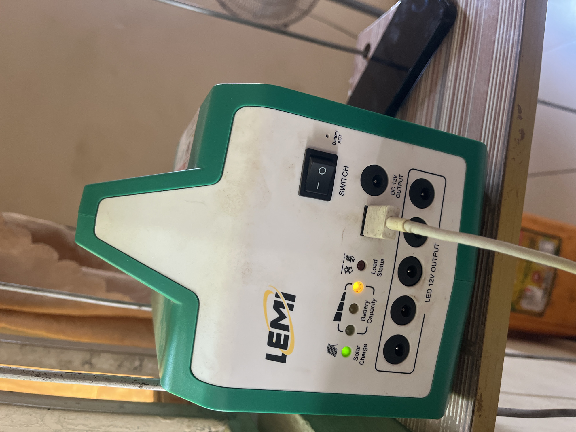 A green and white LEMI brand solar power unit sits on a wooden shelf. The unit has indicator lights for "Solar Charge," "Battery Capacity," and "Load Status." The "Solar Charge" light is green, and the "Load Status" light is yellow. There is a black ON/OFF switch labeled "SWITCH" and a small light labeled "Battery ACT." Below these are four circular ports labeled "LED 12V OUTPUT." A white cable is plugged into one of these ports. In the background, vertical metal bars and orange curtains are visible. A black smartphone rests on the shelf to the right of the solar unit. This image was captured in Jalingo, Nigeria.
