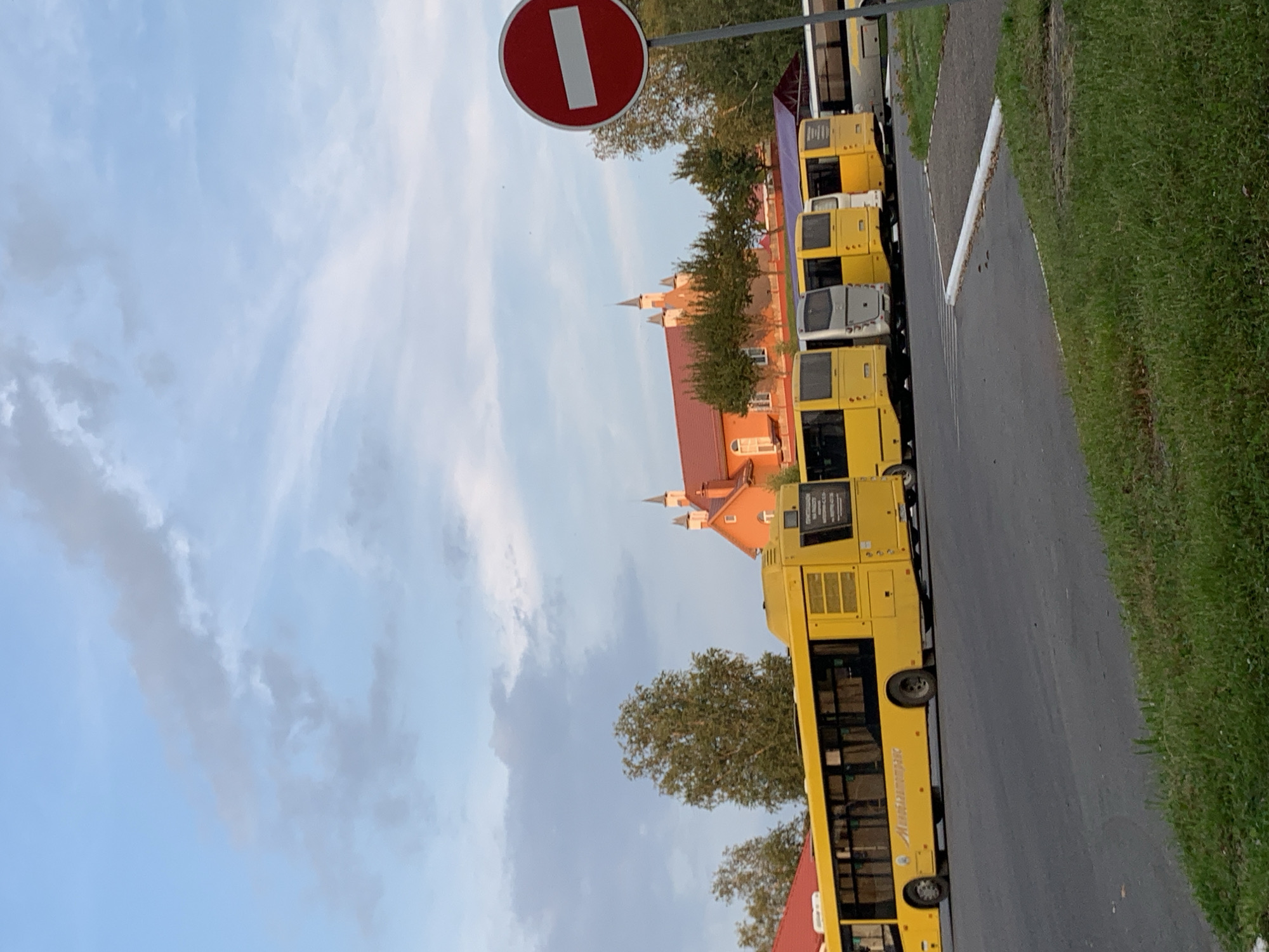 The image captures a view of a street in Kapyĺ, Belarus, with several yellow buses parked in a row. In the background, an orange building with a red roof and decorative turrets is visible, along with several trees. A red circular traffic sign with a white horizontal bar, indicating "no entry," is positioned on the right side of the frame. The sky is partly cloudy, with a mix of blue and gray clouds. The foreground features a grassy area and an asphalt road. There are no people visible in the image. The scene appears to be an outdoor parking area or bus depot.