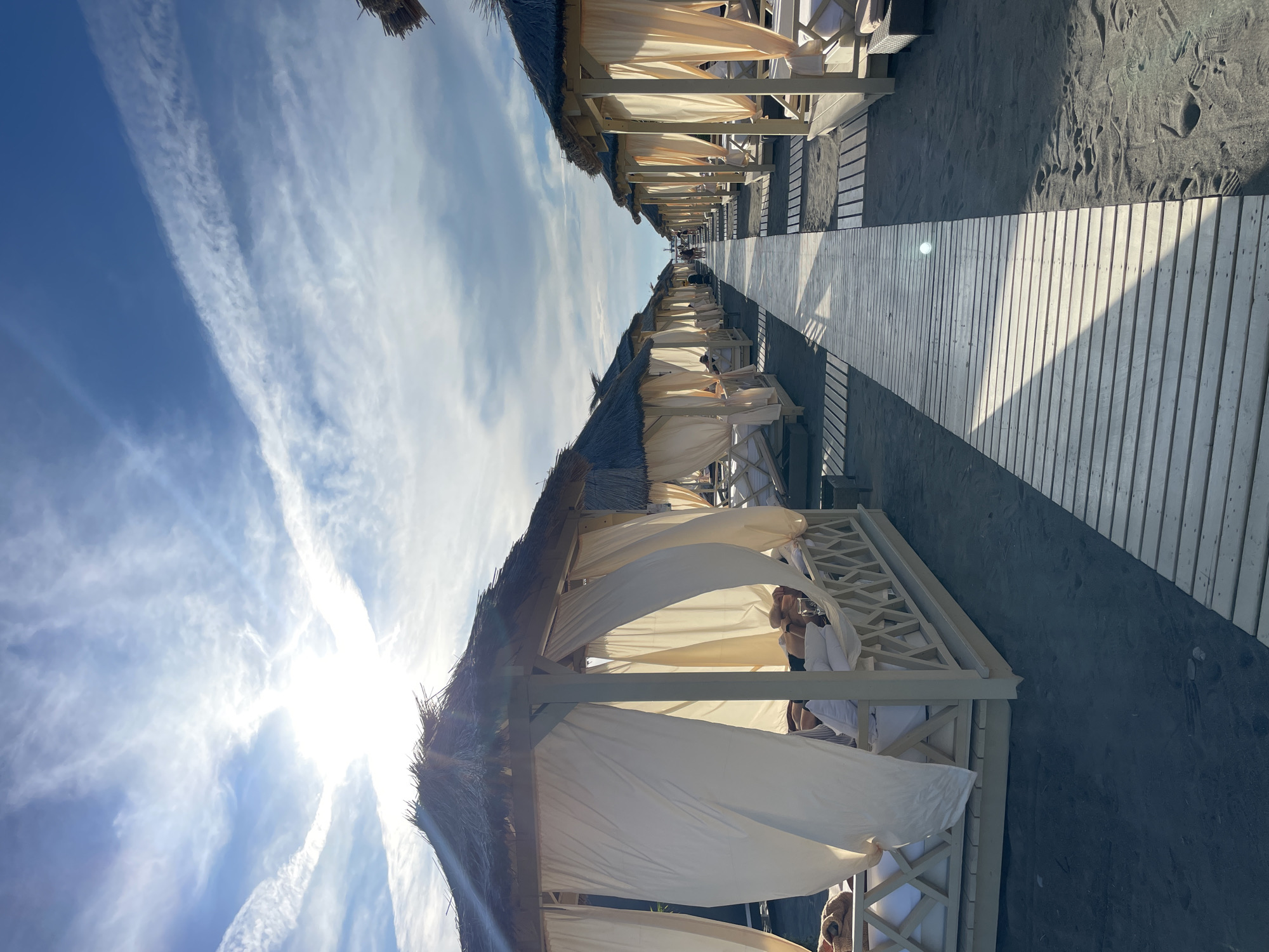 The image depicts a serene beach scene in Sochi, Russia, characterized by rows of luxury cabanas set up on a dark sand beach under a bright, partly cloudy sky. The cabanas are white with thatched roofs and are adorned with flowing cream-colored drapes, creating a sense of privacy and relaxation. Each cabana features a raised bed with white cushions, and several appear occupied by people lounging, some appearing to be in white robes, suggesting a spa-like or resort setting. A wooden boardwalk stretches along the beach, providing a clear path between the cabanas. The sun is visible in the upper left quadrant of the image, casting a bright light and creating a gentle glare. The overall impression is one of tranquility and upscale beachside leisure.