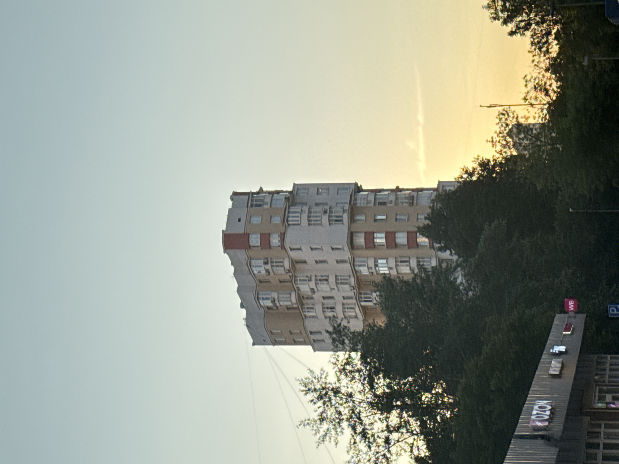 This media file depicts an outdoor scene in Moscow, Russia, during what appears to be either sunrise or sunset, given the warm, hazy glow in the sky. The primary subject is a multi-story apartment building with a cream and reddish-brown facade, featuring numerous windows and balconies. The building is partially obscured by lush green trees, indicating a time of year with full foliage. In the foreground, to the right, is a distinctive outdoor structure with several illuminated signs, one of which clearly displays "OZON" in white lettering against a dark background. Another sign with "WB" in white on a red background is also visible, along with a blue parking sign. The overall impression is a quiet urban moment, with the natural elements of trees and sky juxtaposed against the man-made structures. There are no people visible in the frame, and the atmosphere feels peaceful, bathed in the soft light of the transitional hours of the day.