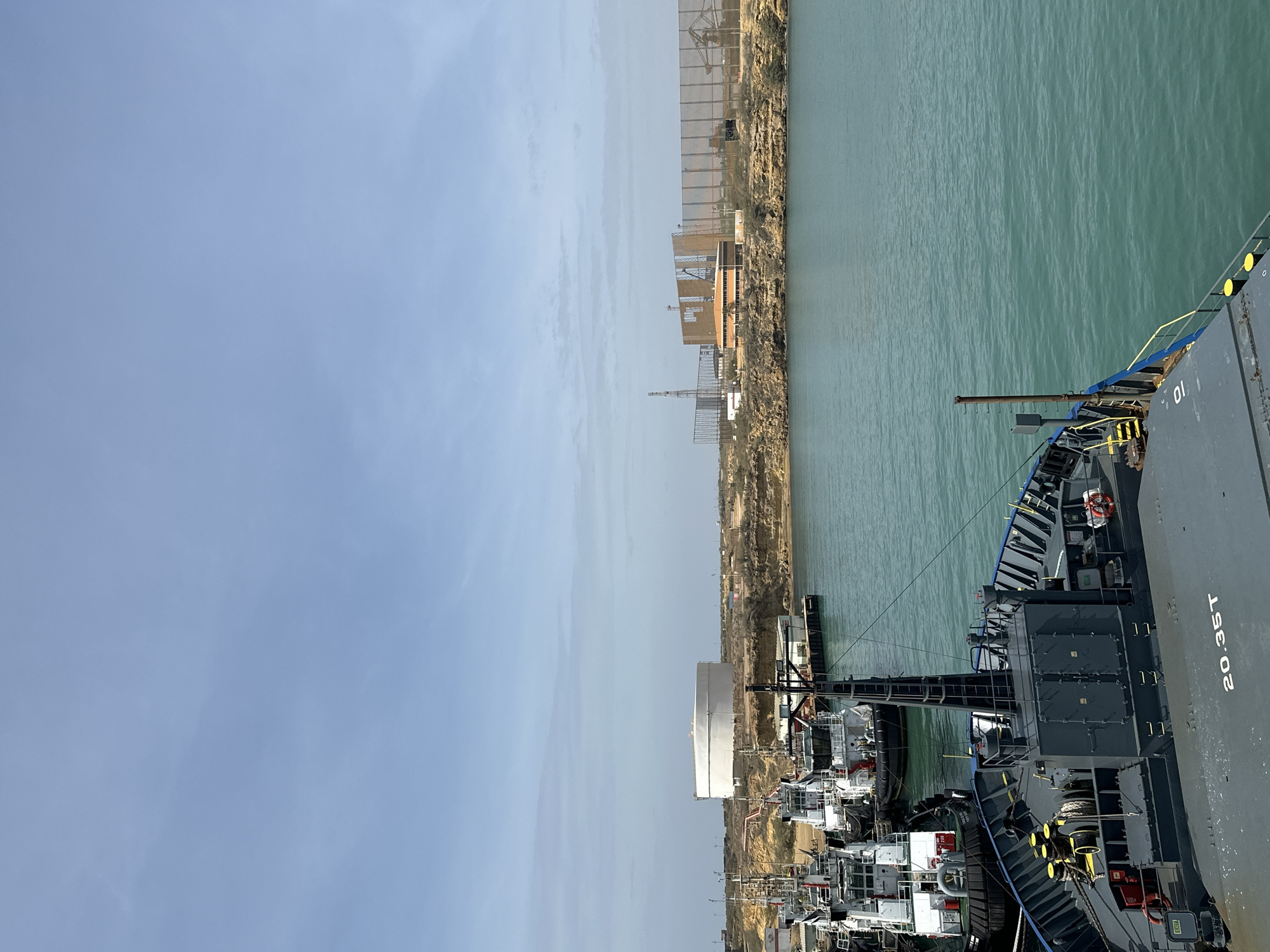 This image depicts a daytime scene at a port or along a canal, featuring a large vessel and an industrial shoreline under a clear sky. The photo is oriented vertically, but when viewed horizontally, a grey vessel dominates the right side, with its bow pointing towards the left.
The vessel, possibly a patrol boat or naval support ship, is dark grey with blue and yellow railings. Visible text on its side includes "01" and "20.35T". A small cannon or gun protrudes from its foredeck, pointed towards the water. Yellow capstans or winches are present on the deck, along with various hatches and equipment, including an orange and white life preserver. Another smaller vessel appears docked directly to the left of the main vessel, and other white utility boats or tugs are visible further along the pier.
To the left of the vessels is a calm body of greenish-blue water, showing slight ripples. Across this waterway, on the left side of the frame, lies a dry, arid shoreline. This landmass is characterized by light brown, rocky terrain. Numerous industrial structures are scattered across the land, including a large, white cylindrical storage tank, metal fences, scaffolding from what appears to be ongoing construction, and other smaller buildings and towers.
The sky above is bright and mostly clear, with a few faint clouds, indicating fair weather and clear visibility during daylight hours. No people are discernible in the image. The overall setting suggests a functional industrial port or transit area, consistent with coastal activities in an arid region like Uribia, Colombia.