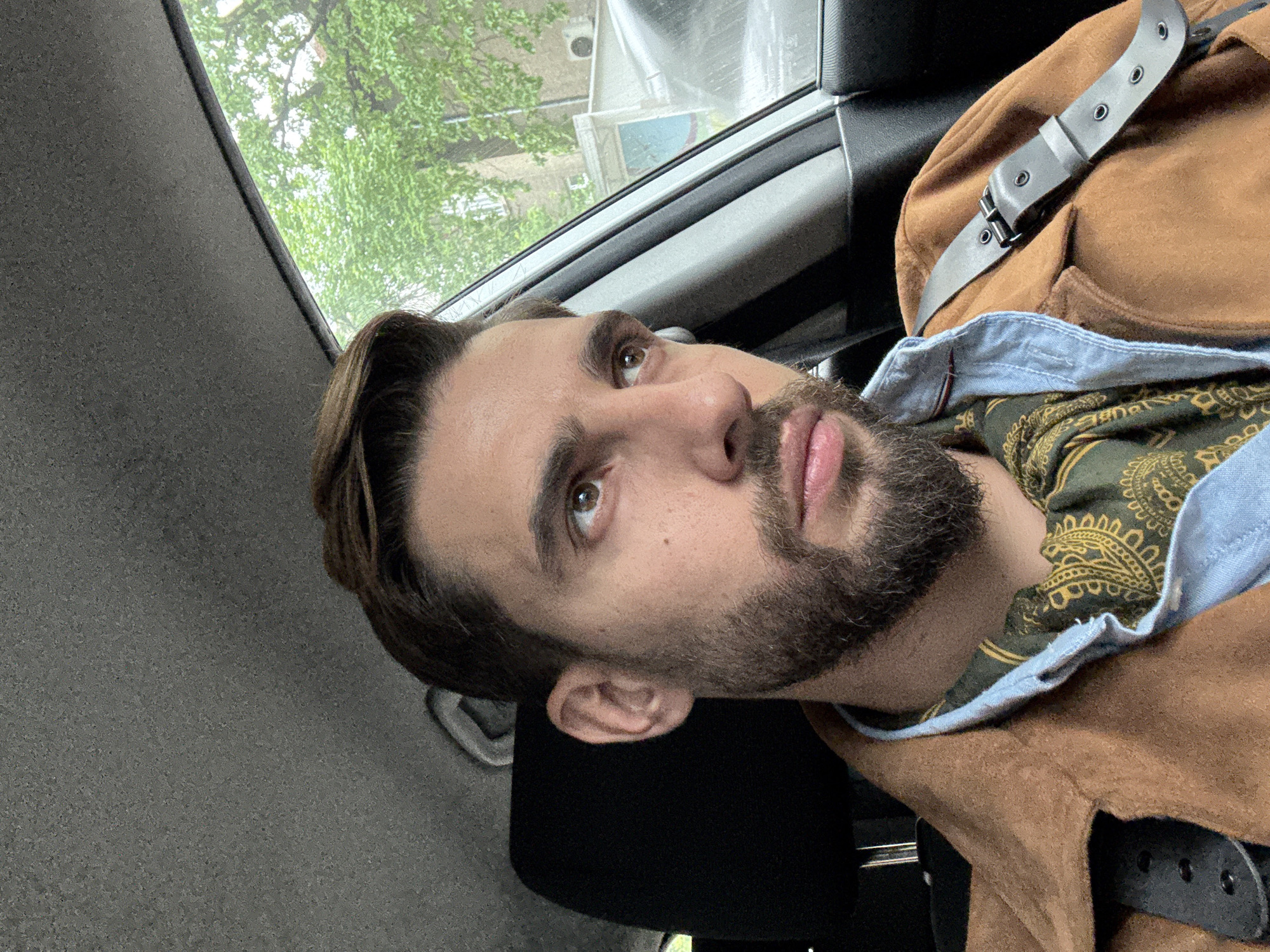 A man with a beard is sitting in a car. He is wearing a brown jacket over a green patterned shirt with a light blue collar. A black leather strap with a metal buckle is draped over his shoulder. Outside the car window, green trees and a building can be seen. Based on the architecture, the location is likely Sofia, Bulgaria.