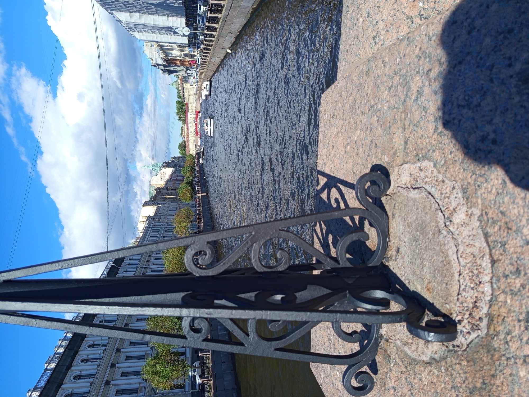 The image shows a canal in Saint Petersburg, Russia, with boats on the water and buildings lining the banks. A stone bridge railing with ornate black metalwork is in the foreground. The sky is blue with fluffy white clouds. The sun appears to be shining, suggesting it is daytime. The canal is calm with gentle ripples. On one side of the canal, there are several boats, including what appear to be tour boats. The buildings on the canal's edge are tall and multi-story, with a classic European architectural style, featuring many windows and decorative elements. Trees with green foliage are visible along the embankment. The overall scene evokes a pleasant, urban atmosphere with a sense of history. The sunlight casts shadows from the railing onto the stone. No discernible text is visible in the image.