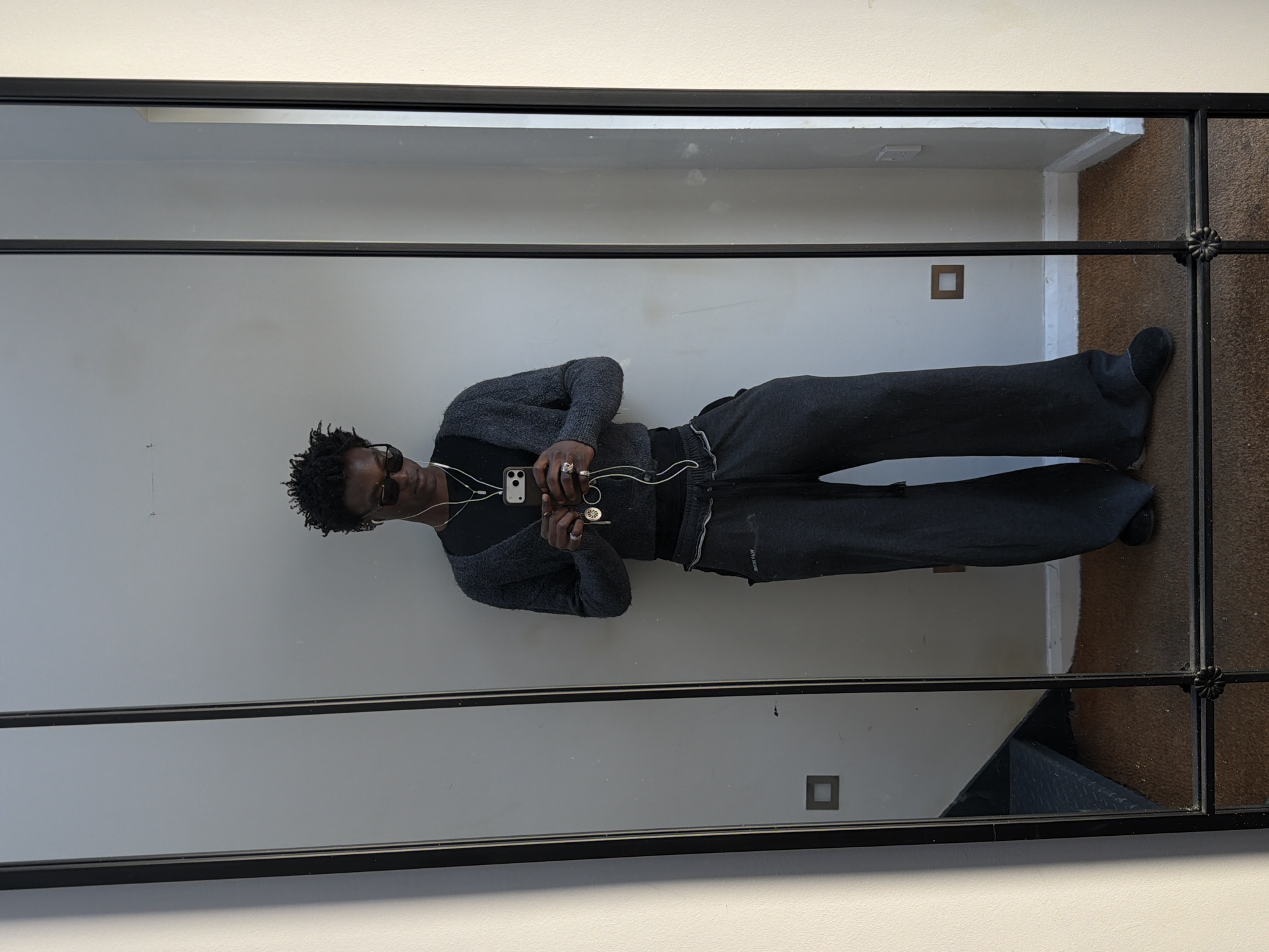 A young person, appearing male, is captured in a mirror selfie taken indoors, likely in London, United Kingdom. The individual stands upright (though the original image is rotated 90 degrees), facing their reflection. They have dark, textured hair styled in short dreadlocks and are wearing dark sunglasses. Their attire consists of a dark grey, subtly textured long-sleeved top and loose-fitting, dark grey trousers, paired with dark footwear. White wired earbuds are visible, with the cord dangling.
The person is holding a black smartphone (likely an iPhone due to its design) in front of them, actively taking the mirror selfie. Several rings are visible on their fingers. On the upper left thigh of their trousers, a small, reversed white text or logo is partially visible, which appears to spell "ALL SAINTS."
The mirror has a prominent black frame, divided into multiple sections by thin black vertical and horizontal bars. A decorative, floral-like embellishment is present on the right side of the mirror frame. The background reflected in the mirror shows a plain, light-colored wall, a darker floor (possibly carpet), and a glimpse into what appears to be another room or hallway through an opening. Two small, square-shaped insets are visible on the reflected wall, possibly decorative elements or light switches. No specific time of day or emotional state is discernible.