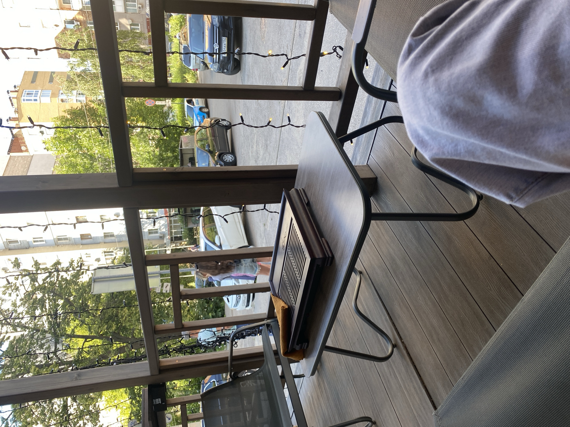 The image shows an outdoor seating area, likely a cafe or restaurant, with a wooden deck and railing. A small, dark wooden table holds a tea tray and a folded yellow cloth. The scene is viewed from behind a chair, with a person's shoulder and arm, wearing a light grey garment, visible in the foreground.
Through the railing, a street scene is visible with cars parked and driving. Buildings with windows and balconies line the street, and trees with green foliage are abundant, suggesting it is daytime and likely a warm season. Small, white lights are strung along the railing, adding a decorative element. The weather appears to be sunny, casting shadows and highlighting the textures of the wooden deck and seating. A sign with Cyrillic text is partially visible on the street. The overall impression is a relaxed outdoor setting in an urban environment.