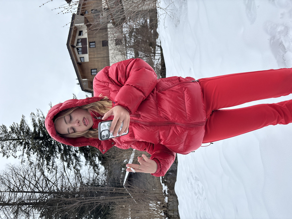 A young individual with blonde hair, wearing a red hooded puffer jacket and matching red pants, stands in a snowy outdoor environment in La Clusaz, France. The individual is centrally positioned, looking downwards at a white smartphone held in their right hand, which has a clear case displaying small green bicycle motifs. A lit cigarette is held between the index and middle fingers of their left hand. The individual's tongue is slightly extended. The ground is covered in white snow. In the background, tall coniferous and bare deciduous trees are visible, along with a multi-story chalet-style building featuring wooden siding, dark window frames, and balconies. The sky is overcast.