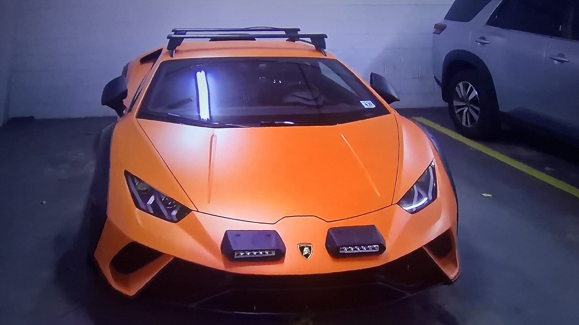 The image shows a matte orange Lamborghini Huracan parked in an indoor parking garage. The Lamborghini has aftermarket accessories, including two light bars mounted on the front hood and a roof rack. The car is angled towards the camera. A silver SUV is parked to the right, partially visible in the frame. A yellow parking line is also visible, delineating parking spaces. The location is Unknown, Unknown.