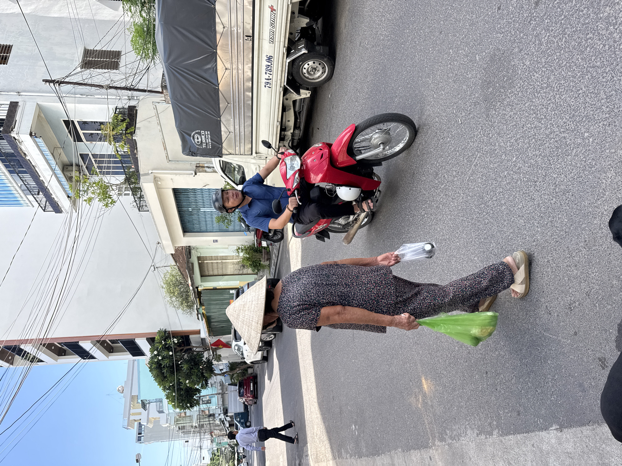 This is a street scene in Nha Trang, Vietnam. A woman wearing a conical hat and a floral patterned outfit walks away from the camera carrying two plastic bags, one green and one clear. She is wearing tan sandals. In the background, a man in a blue collared shirt and dark pants rides a red motorcycle, wearing a black helmet. Behind the motorcycle is a white truck with a black tarp covering its cargo bed. The truck has the license plate number 79A-789.06. The street is paved with asphalt and has various buildings lining its sides. The sky is clear and blue.