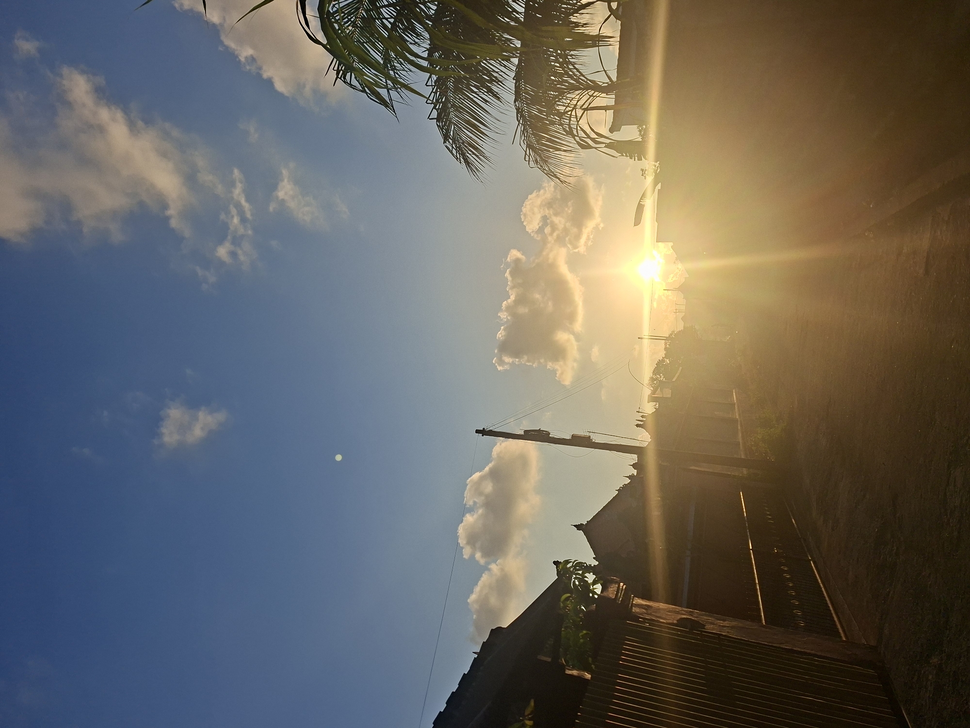 The image depicts a street scene in Blahbatuh, Indonesia, at sunset. The sun is low in the sky, casting a strong golden light that creates lens flares and washes over the scene. To the left, a dark metal fence with vertical slats lines the edge of a path or road. Beyond the fence, the dark roofs of traditional Balinese buildings are visible, with some greenery peeking out. A utility pole with electrical wires stands in the middle ground. On the right side of the image, a large palm tree with prominent fronds is silhouetted against the bright sky. The sky is a clear blue with scattered white and light orange clouds, illuminated by the setting sun. The ground appears to be a dirt or gravel path. No people are visible in the frame.