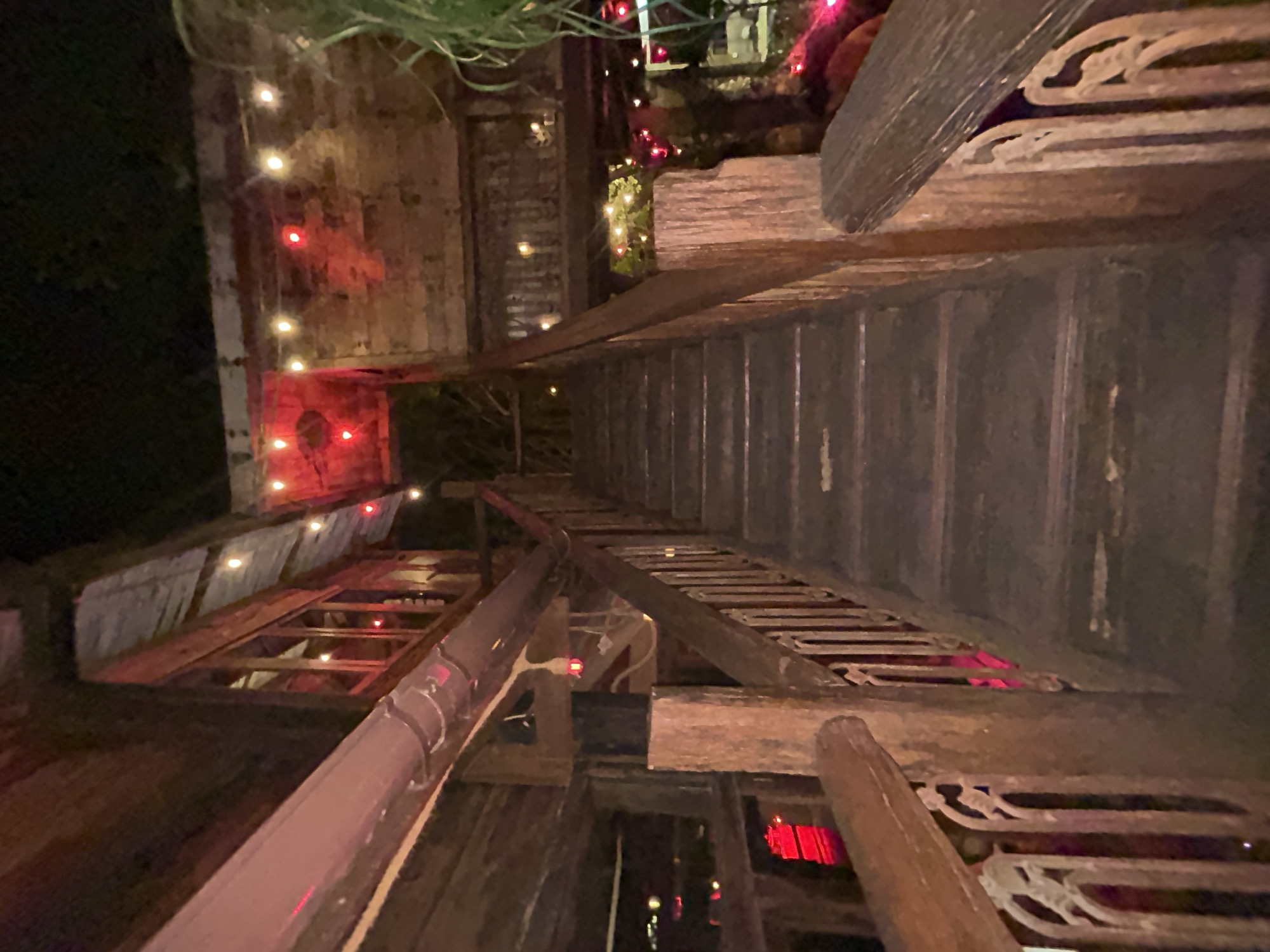 The image depicts an outdoor staircase at night, constructed from weathered dark wood. The staircase is flanked by wooden railings with ornate, possibly metal, balusters. String lights, some red and some yellow, are visible along the upper structures of the building and on the sides of the stairs, casting a warm glow and illuminating the scene. The architecture appears rustic and somewhat aged. To the left of the stairs, a large, curved pipe runs horizontally. Red light is visible reflecting from below. The overall impression is of an atmospheric, dimly lit outdoor venue, possibly a restaurant or bar. The location is specified as Mengwi, Indonesia.
