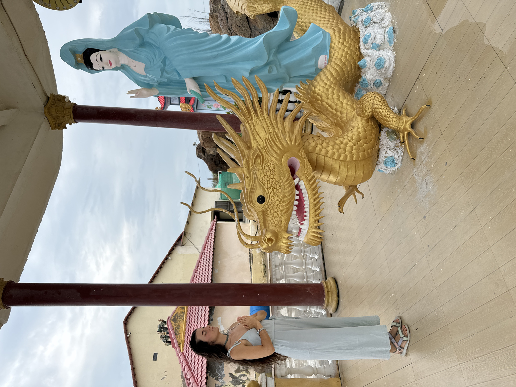 A woman in a light blue dress and sandals stands with her hands clasped in prayer in front of a golden dragon statue and a light blue statue of a woman. The statues are on an elevated platform, and a column is visible behind the woman. In the background, there is a building with a red tile roof. The image was taken in Mui Ne, Vietnam.
