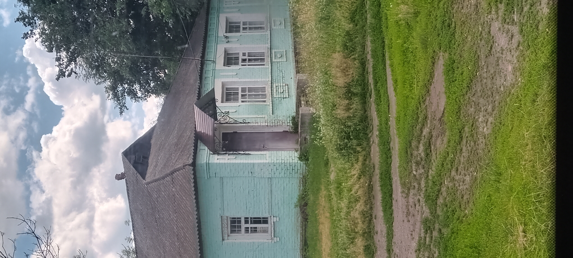 A light blue brick house with a dark grey roof stands in the city of Koviahy, Ukraine. The house has a prominent entrance with a decorative awning. Several windows with white trim are visible along the front of the house. The building is surrounded by green grass and some overgrown vegetation. The sky above is partly cloudy with white and grey clouds. The scene depicts a residential dwelling in a rural or semi-rural setting.
