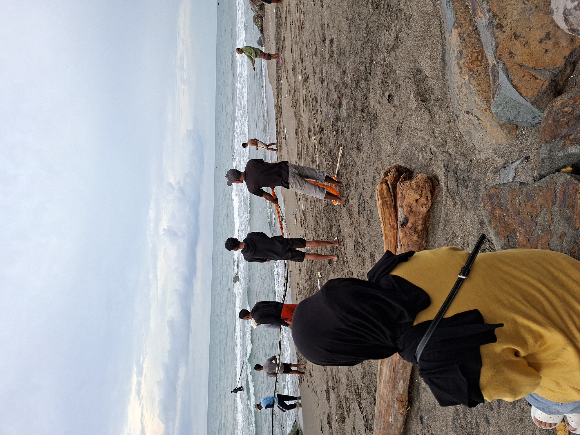 This media file captures a coastal scene with several people engaged in activity on a sandy beach. The image is composed with an expansive, light grey, overcast sky dominating the left side, meeting the ocean horizon.
In the foreground, positioned on the right, a person is seen from behind, wearing a bright yellow long-sleeved top, a black head covering (possibly a hijab), and light blue jeans, with white sandals visible on their feet. A black strap, likely from a bag, crosses their back. This individual appears to be observing the scene unfolding towards the ocean. Adjacent to this person are large, textured rocks and weathered driftwood pieces scattered on the dark sand.
Along the water's edge, stretching across the middle ground, are several men. Their primary activity appears to be fishing or pulling nets from the ocean. One man, prominently featured in a black t-shirt and grey shorts, is actively pulling an orange net or rope from the breaking waves. Further down the shore, other men are seen standing near the water; one in a black hoodie and shorts, another in a green shirt and brown shorts, and others holding long poles that could be fishing rods or implements for managing nets. Some figures are partially obscured or in the distance.
The ocean on the right side of the frame shows gentle waves breaking on the dark, damp sand. The rocks on the right are varied in color, with shades of grey, brown, and orange, suggesting an ancient geological formation or breakwater. The overall lighting is soft and diffused due to the cloudy conditions, hinting at either an early morning or late afternoon setting. No discernible text is visible in the image. The scene conveys a sense of communal effort or leisure activity by the sea in Lhokseumawe, Indonesia.