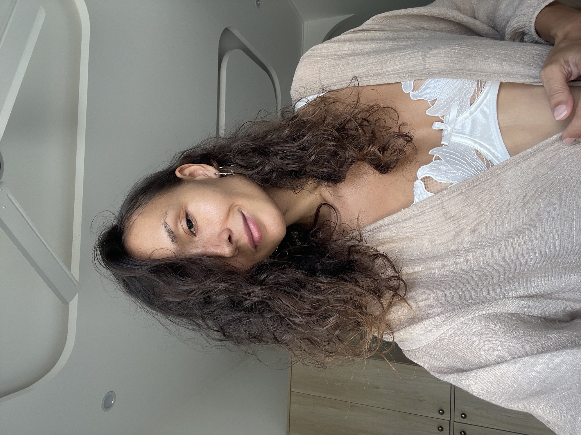 A woman with long, curly brown hair, wearing a white lace bra and a beige open robe, poses for a selfie in a room with light-colored walls and a ceiling with recessed lighting. A wooden closet is visible in the background.