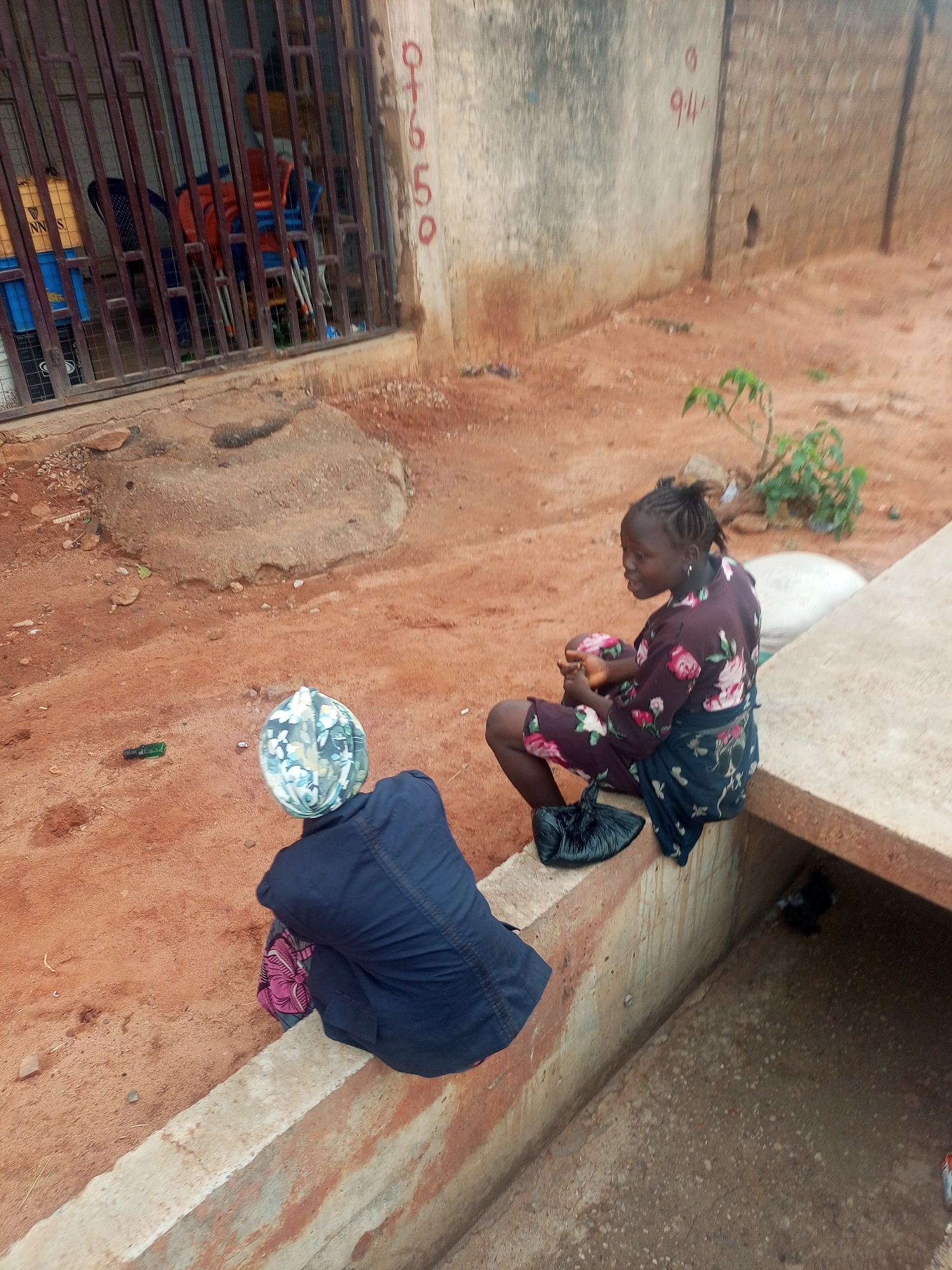 Two women are sitting near a roadside drain in a dusty outdoor environment. One woman faces the viewer with a floral dress. She is sitting on a concrete edge. Next to her is a black plastic bag. The other woman faces away, wearing a dark shirt and a headscarf. To the left, a metal gate with stacks of chairs is visible. In the background, there is a concrete wall with painted numbers "911" and "07650". The ground is reddish-brown with scattered debris and vegetation.