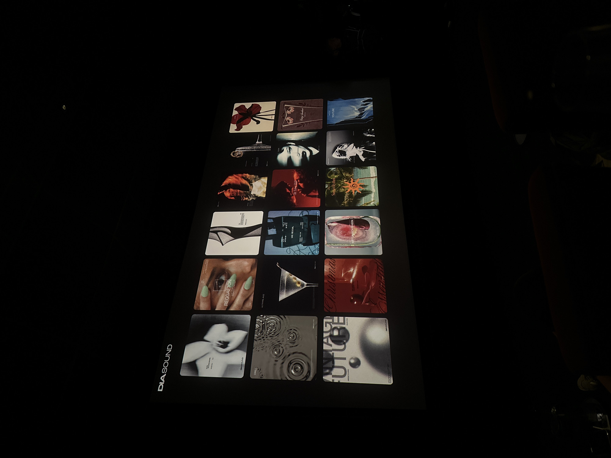 Here is a forensic description of the image:
The image shows a tabletop display in a dimly lit environment. The display features a grid of eighteen square images, each depicting various abstract or artistic themes. A logo "DIASOUND" is visible on the bottom left corner of the display. The images on the display vary in color and content, including floral motifs, portraits, landscapes, and abstract designs. Text labels are present under some of the images, with words like "DRILL", "REGGAETON", "ULTAMIE JAZZ", "UNS REVIVAL", "Sunset Rythm", "YOUTUBE" visible. The overall scene suggests a presentation or display of artistic works, possibly related to music or sound design. The location cannot be determined from this image.