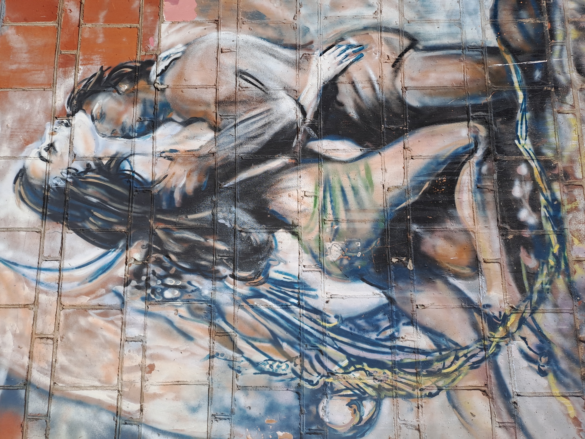 The image shows a street art mural painted on a brick wall in Elektrostal, Russia. The mural depicts a man and a woman in an embrace, possibly a kiss. The figures are rendered in shades of gray, black, blue, and tan, with visible brushstrokes. The woman has blue fingernails. A tangle of blue and yellow lines surrounds their bodies. The background is the brick wall itself, with patches of orange and pink paint.
