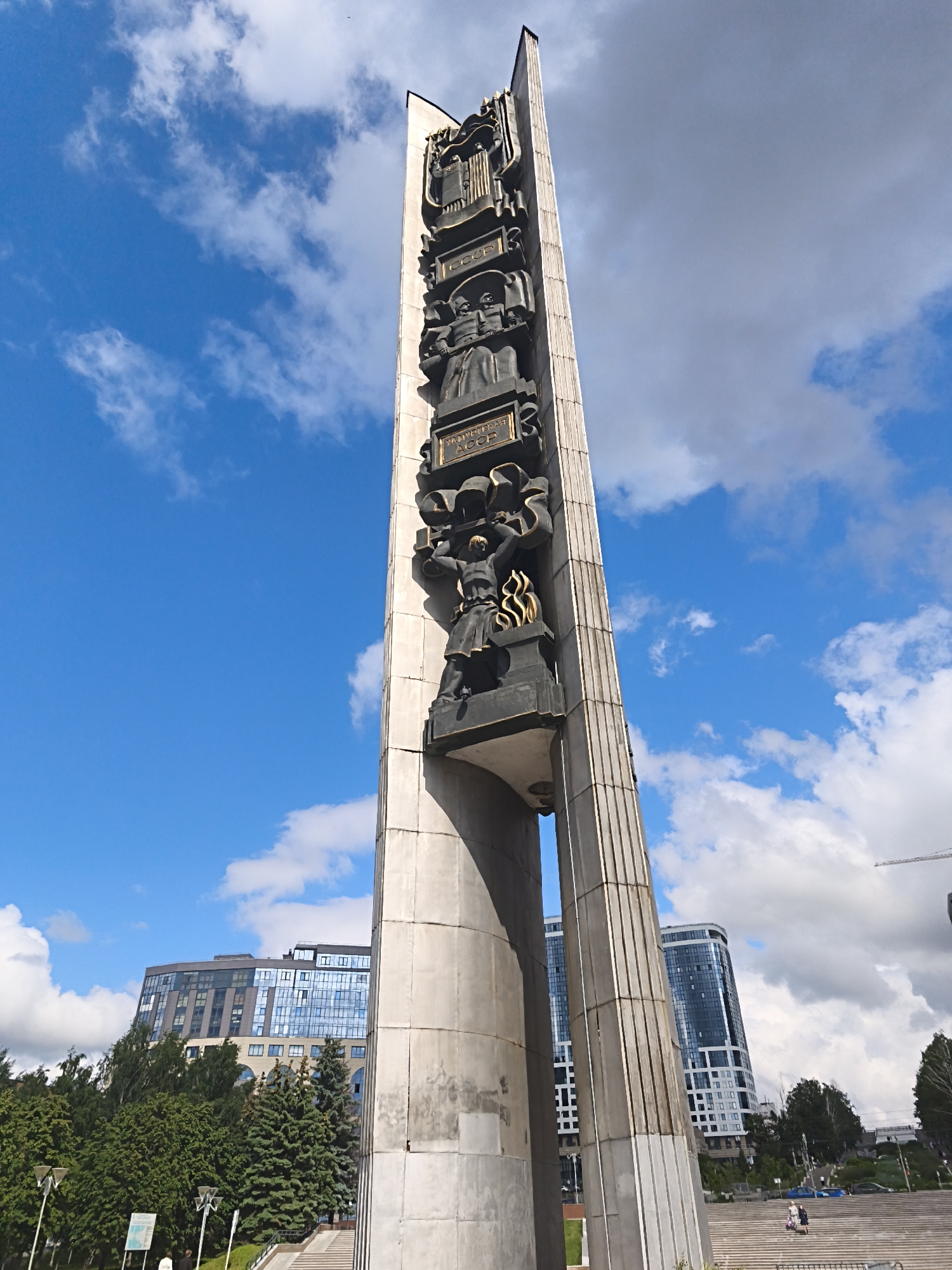 A tall, monumental stele or obelisk, likely made of light-colored stone or concrete, dominates the scene under a bright blue sky with scattered white and some darker grey clouds. The monument features multiple large, dark bronze relief sculptures stacked vertically on its front face. These reliefs depict various figures and symbols, including what appears to be an open book or scrolls at the top, followed by figures in formal attire, and further down, figures possibly representing workers or soldiers. One prominent figure at the bottom section holds a torch or flame.
Several plaques with visible Cyrillic text are integrated into the reliefs. "СССР" (USSR) is clearly legible multiple times, as is "УДМУРТСКАЯ АССР" (Udmurt ASSR) on a lower plaque, indicating the monument's historical context.
The monument is set in an urban environment, with modern, multi-story buildings featuring glass facades visible in the background, particularly on the right side. Lush green trees, including both conifers and deciduous types, line the base of the monument and the surrounding area, suggesting a park or public square. A wide set of concrete or stone steps leads up towards the monument from the lower right foreground, where a few people can be seen walking. Lamp posts and a sign are visible in the lower left. The time of day is bright daylight, and a construction crane is visible in the distant background on the right. The overall setting is a public space, characteristic of a memorial or historical landmark in an urban Russian city like Izhevsk.