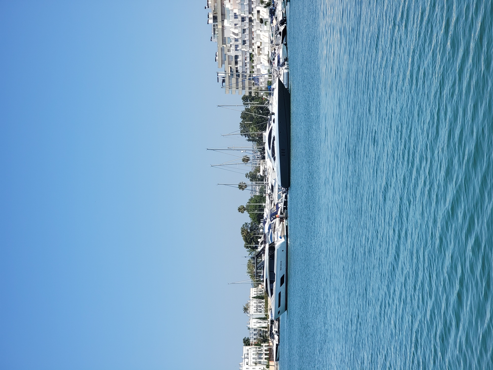 A wide-angle, eye-level, vertical shot captures a marina in Quarteira, Portugal, on a clear, sunny day under a bright blue sky. The foreground is dominated by calm, blue water with gentle ripples. In the midground, several luxury yachts and sailboats are docked. Two prominent yachts, one white with dark trim and another dark gray, are positioned centrally. Behind the yachts, a row of white apartment buildings with balconies and a hotel-like structure are visible, interspersed with green trees and palm trees. The scene depicts a serene harbor environment with modern architecture and vessels.