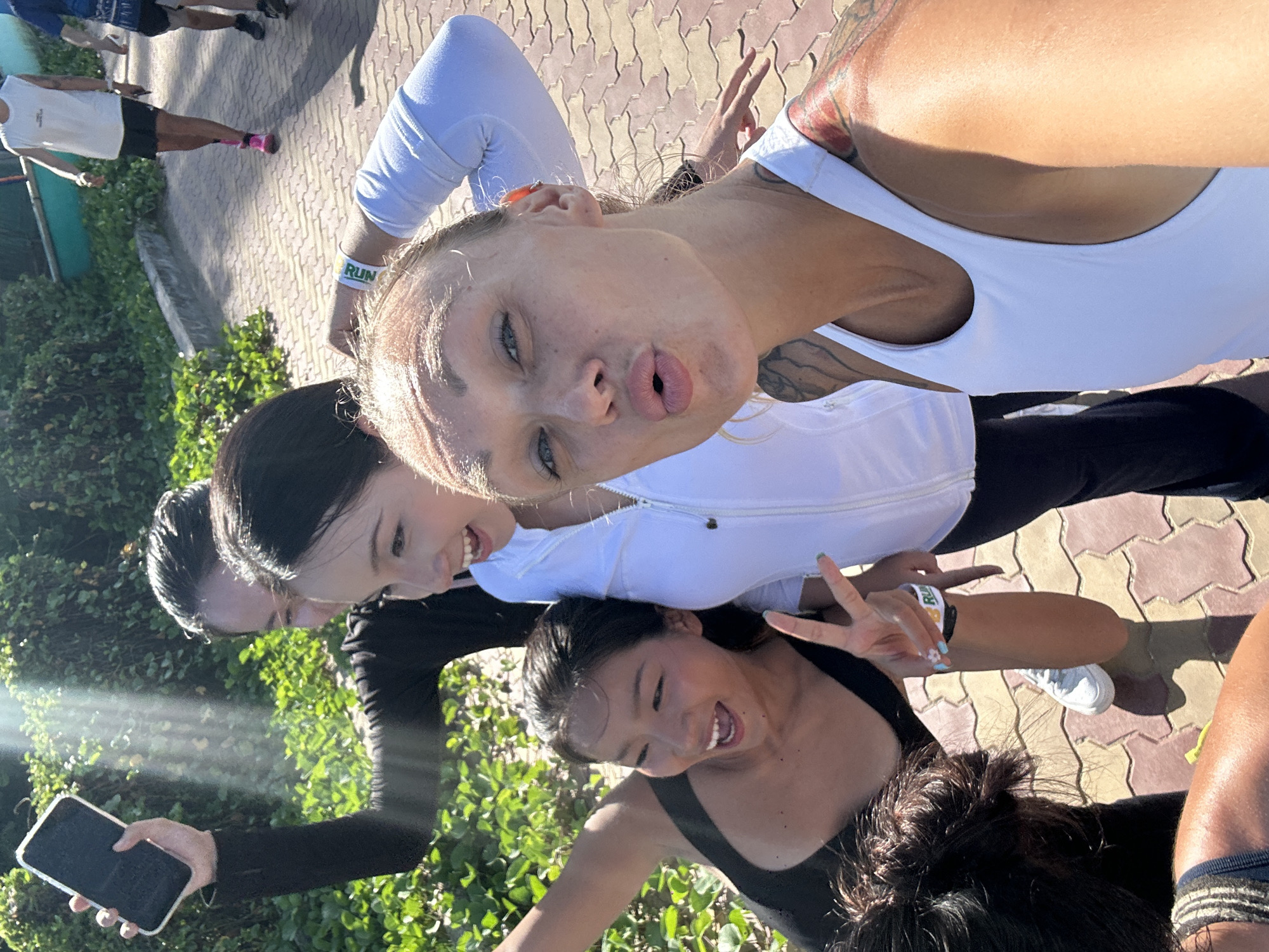 In Mũi Né, Vietnam, on a sun-drenched, paved walkway, five individuals are captured in a candid group selfie. The foreground is dominated by a woman with blonde hair, wearing a white tank top, making a duck face and holding a phone. To her left, a woman in a black tank top, with dark hair, smiles and makes a peace sign with her hand, her nails painted with a light blue polish and tiny white flowers. Behind them, another woman in a white zip-up jacket grins widely. Further back, another individual in a black long-sleeved shirt holds a phone aloft, seemingly taking the picture. In the background, two men in athletic wear walk away, one in a white sleeveless shirt and black shorts, the other in a blue shirt and black shorts. Lush green foliage lines the walkway on the left, with a glimpse of a turquoise structure in the distance on the right, suggesting a coastal or resort-like setting. The pavement is made of interlocking red and beige hexagonal tiles. Sunlight streams in from the upper left, casting dappled shadows and highlighting the scene.