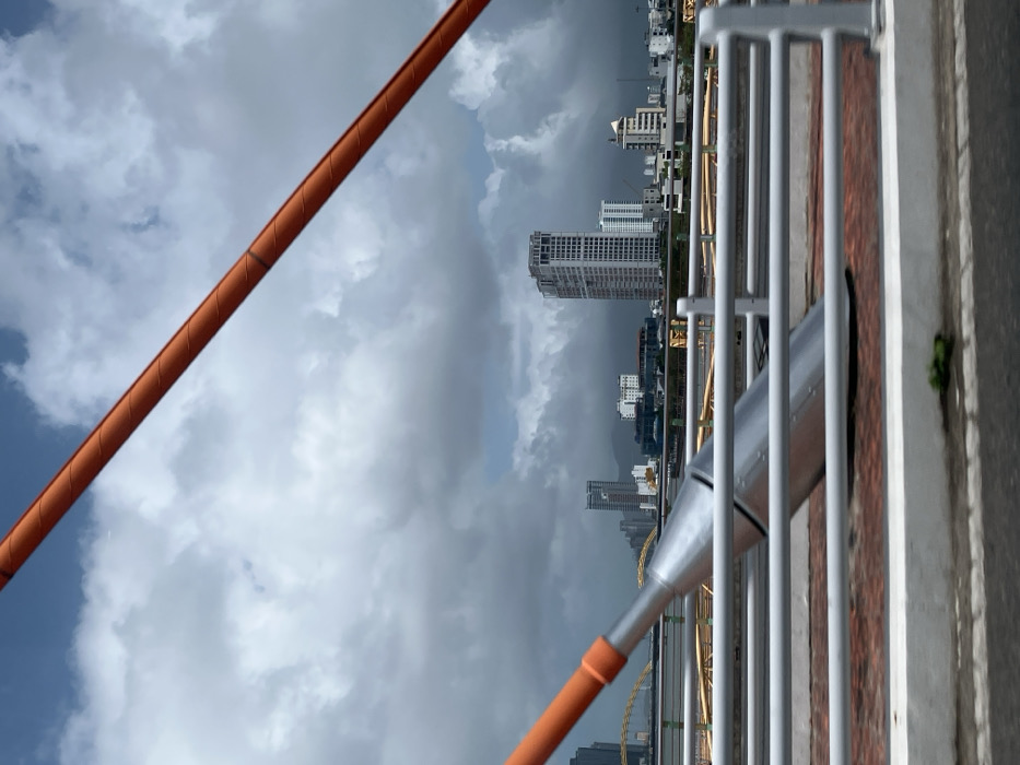 The image depicts an urban cityscape viewed from a cable-stayed bridge, specifically located in Da Nang, Vietnam, within the area providing views of Ngũ Hành Sơn Ward. The perspective is outward from the bridge, showing a river and a city skyline under a heavily clouded sky with some patches of blue.
In the foreground, two bright orange, spirally textured bridge cables diagonally cross the frame: one from the upper right extending downward, and another entering from the lower left. Below the upper-right cable, a large, silver-grey, conical-cylindrical metallic component, approximately two meters in visible length, angles downwards from the left, partially obscured by a light-grey multi-bar horizontal guardrail system. This guardrail is mounted on a reddish-brown textured surface, likely a pedestrian or cycle path, which is bordered by a white concrete curb. A dark asphalt road surface occupies the very bottom edge of the frame.
Across a body of water, the midground presents a dense urban skyline featuring numerous high-rise buildings and skyscrapers. A prominent tall, light-colored building with multiple windows dominates the central view. To the left, a section of a bridge characterized by yellow/gold arched supports is discernible (identified as the Dragon Bridge). Distant green mountains or hills form the horizon line behind the city structures. No individuals or specific actions are visible in the scene. The overall lighting suggests an overcast day.