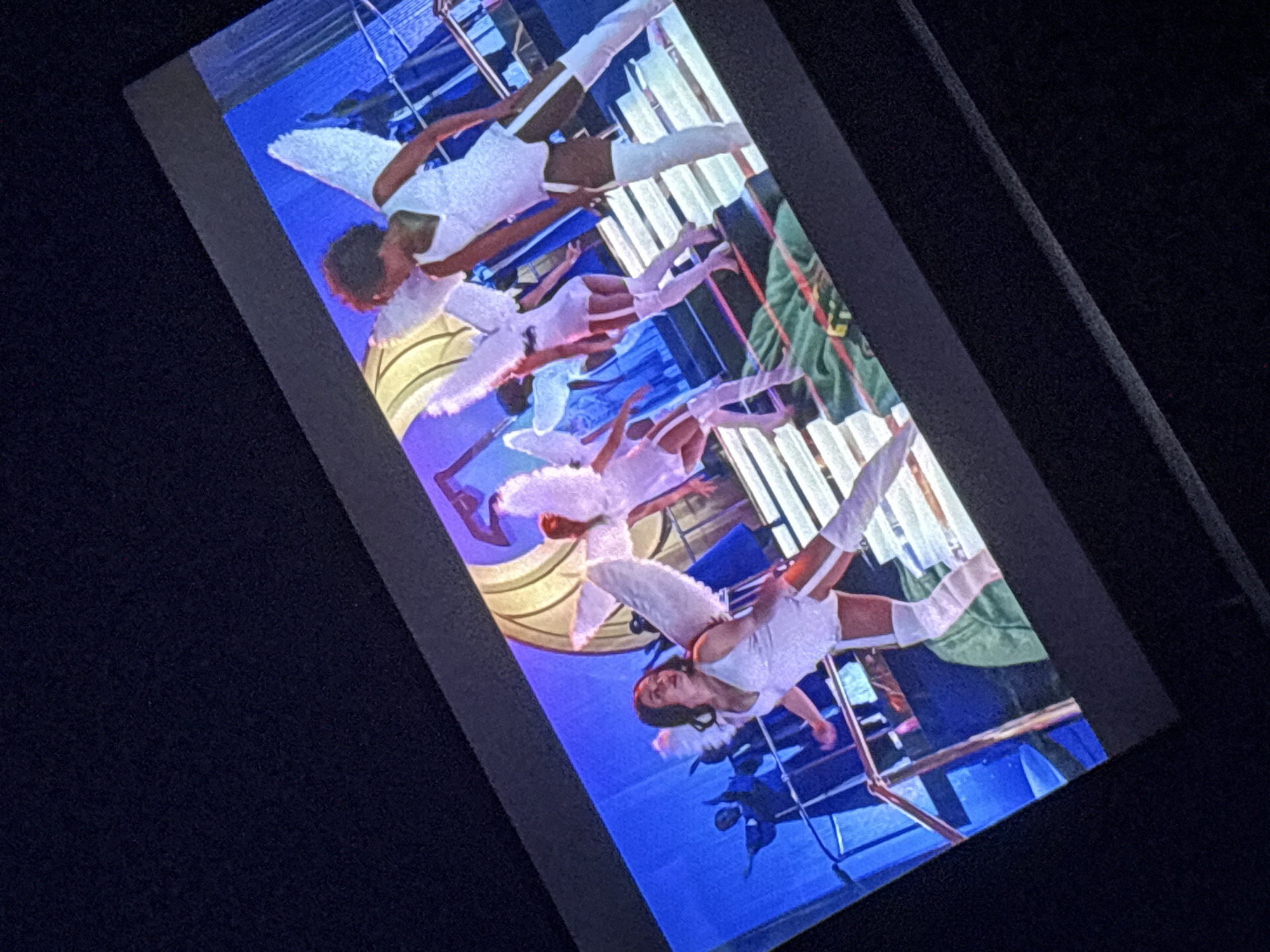 The image displays a screen showing a performance with several individuals dressed as angels. These performers are wearing white costumes that include what appear to be wings. The scene is set on a stage with blue lighting and a backdrop featuring a large, stylized gold structure. The performance appears to be taking place indoors on a stage. The location in the city of Dnipro, Ukraine, cannot be determined from the provided image.