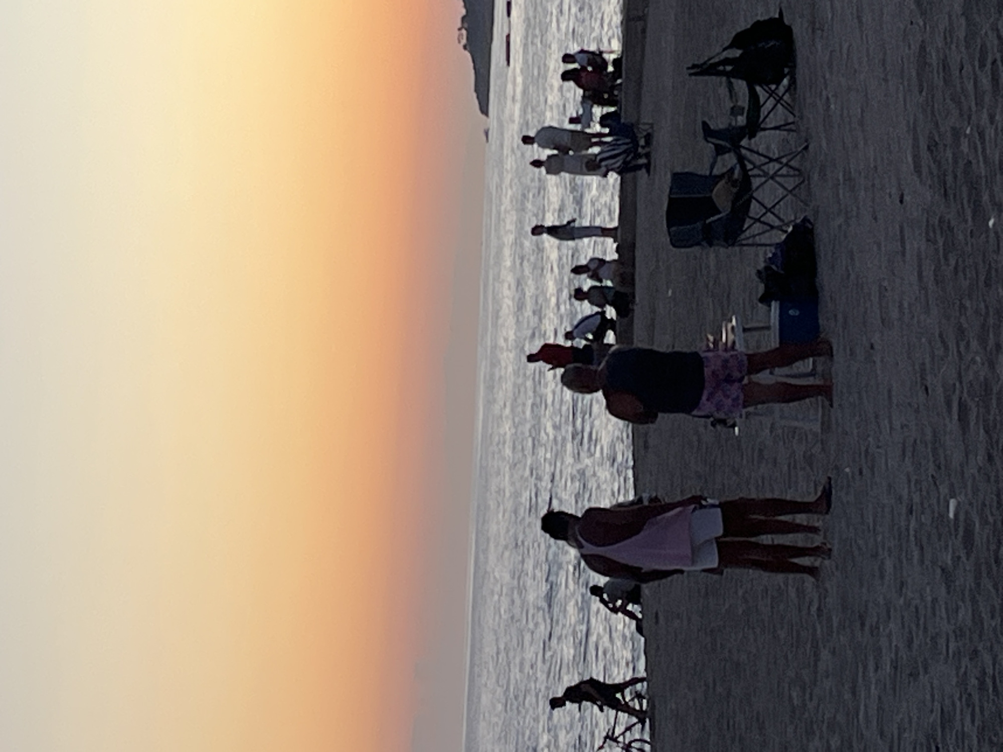 The image shows a beach scene at sunset. The sky transitions from pale yellow to orange towards the horizon. A body of water, presumably the sea, reflects the colors of the sky. Along the shoreline, several people are visible, some standing and some sitting. There are beach chairs present. At least one person is riding a bicycle. The image was taken at Alanya, Turkey.