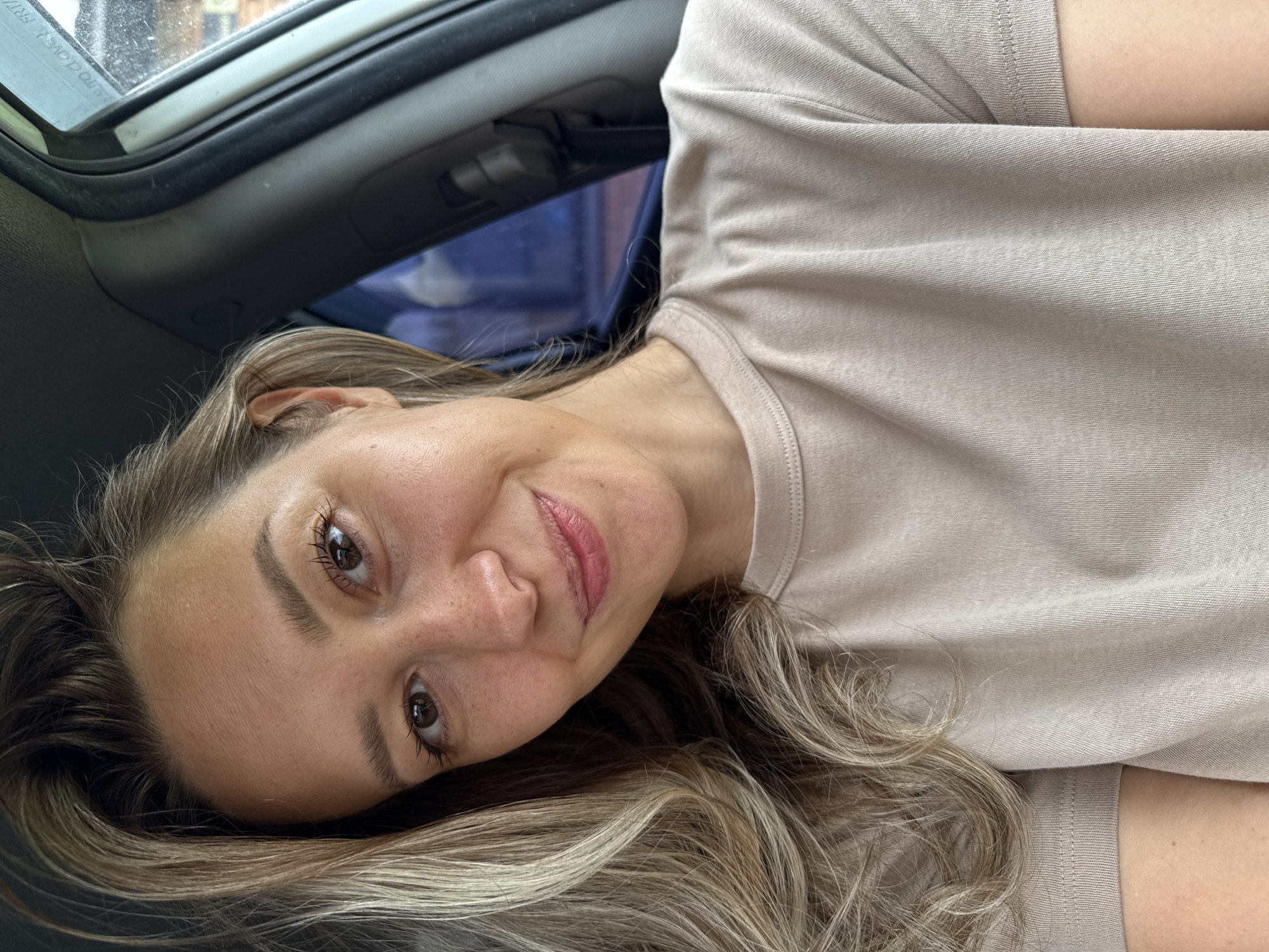 An unidentifiable woman is seen from the chest up, inside a vehicle. She has blonde hair with darker roots, and her hair is styled in loose waves that fall over her left shoulder and across her chest. Her skin is light-toned with visible freckles on her nose and cheeks. She is wearing a light beige t-shirt with a crew neck and short sleeves. Her head is tilted slightly to her left, and she is smiling gently at the camera. Her eyes are brown and her eyelashes are dark. The background shows the interior of a car, with a dark gray dashboard and a window with a blue tint. Some smudges and dust are visible on the car's window frame. The image appears to have been taken during the daytime. There is no information available to determine the location within the city of Chaikovsky, Russia.