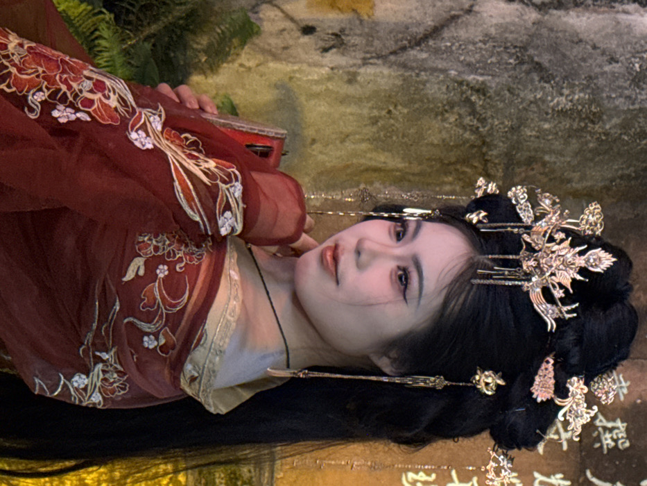 A young East Asian female is depicted in a three-quarter profile against a textured wall in Yuzhong District, China. She wears an elaborate traditional Chinese-style red garment with gold and white floral embroidery. Her dark hair is styled in an intricate updo, featuring a prominent gold-colored phoenix-motif headpiece with multiple dangling ornaments. Her face exhibits a slight smile, with her gaze directed towards the left, accentuated by visible makeup including eyeliner and reddish-orange lipstick. Her right hand is gently positioned near her chin, while her left hand holds a red object, likely a smartphone, against her chest. A thin dark necklace is visible around her neck. The background consists of a mottled, distressed wall showing shades of grey, brown, and yellow-green, with faded Chinese characters partially visible on the right. Dim, warm lighting illuminates the scene, and some dark green foliage is present in the lower-left corner.