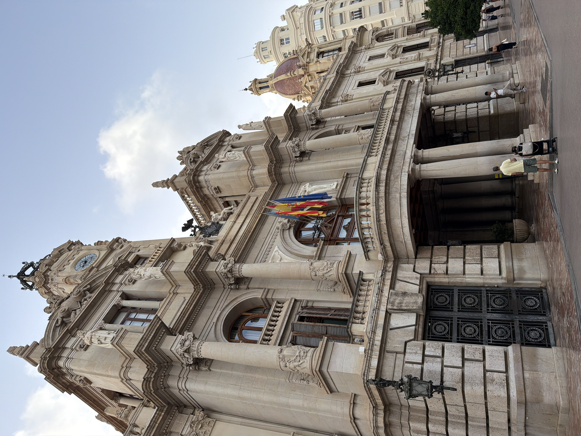The image shows a low-angle, wide shot of a grand, ornate building with intricate stonework. The building features multiple stories, balconies, columns, statues, and a prominent clock tower with a circular clock face. Several flags, including the Spanish flag, are displayed prominently on a balcony. In the foreground, there are several people walking on a paved area in front of the building. The sky above is a clear blue with a few white clouds. The lighting suggests it is daytime, likely late afternoon. The architectural style appears to be classical or baroque.