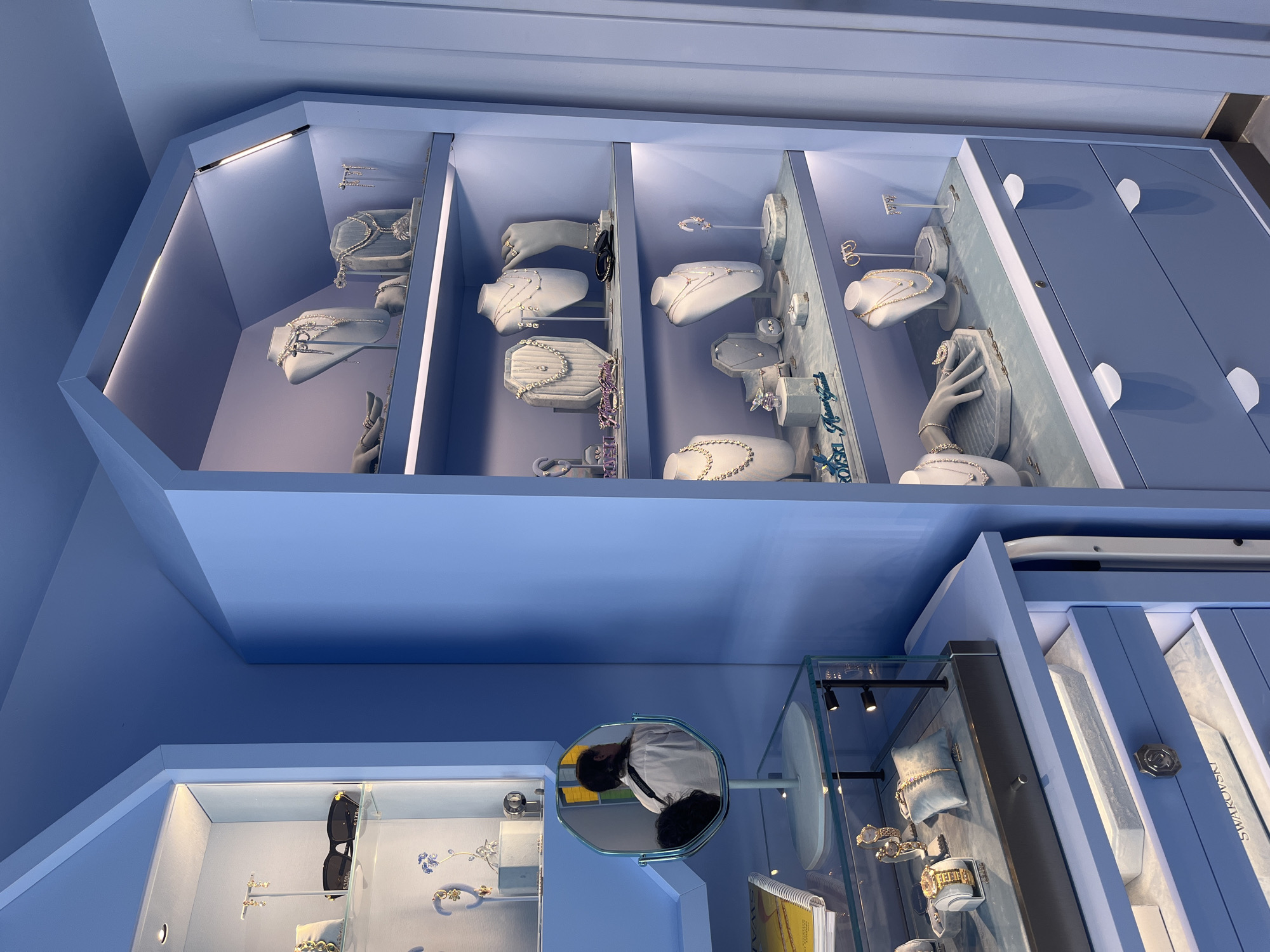 The image shows a bright blue retail display of jewelry in what appears to be a store. The display features multiple shelves filled with necklaces, earrings, bracelets, and rings. Some items are showcased on mannequins and stands, while others are placed within glass cases. The lighting in the display is soft and focused, highlighting the products. In the foreground, a mirror reflects a person wearing a white shirt, facing away from the camera. The overall aesthetic is modern and elegant, with a clean and organized presentation of the jewelry. The store appears to be located in Budva, Montenegro.