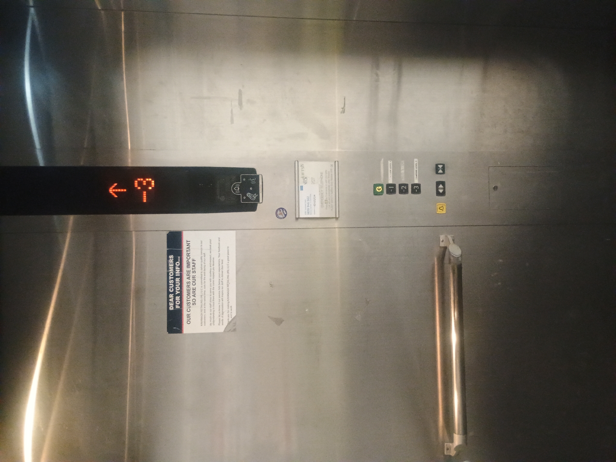 An elevator panel is shown in the image. The elevator is not moving as indicated by the illuminated indicator showing "-3" and an upward arrow. The panel includes buttons for levels G, -1, -2, and -3, with a label "CARPARK LEVEL 3" next to the -3 button. There are also buttons for alarm, door open, and door close. A sign above the buttons reads "DEAR CUSTOMERS FOR YOUR INFO... OUR CUSTOMERS ARE IMPORTANT SO ARE OUR STAFF". Another sign below mentions "EMERGENCY TELEPHONE" and provides contact numbers. There are also two metal handles mounted on the elevator wall. The elevator appears to be made of stainless steel.