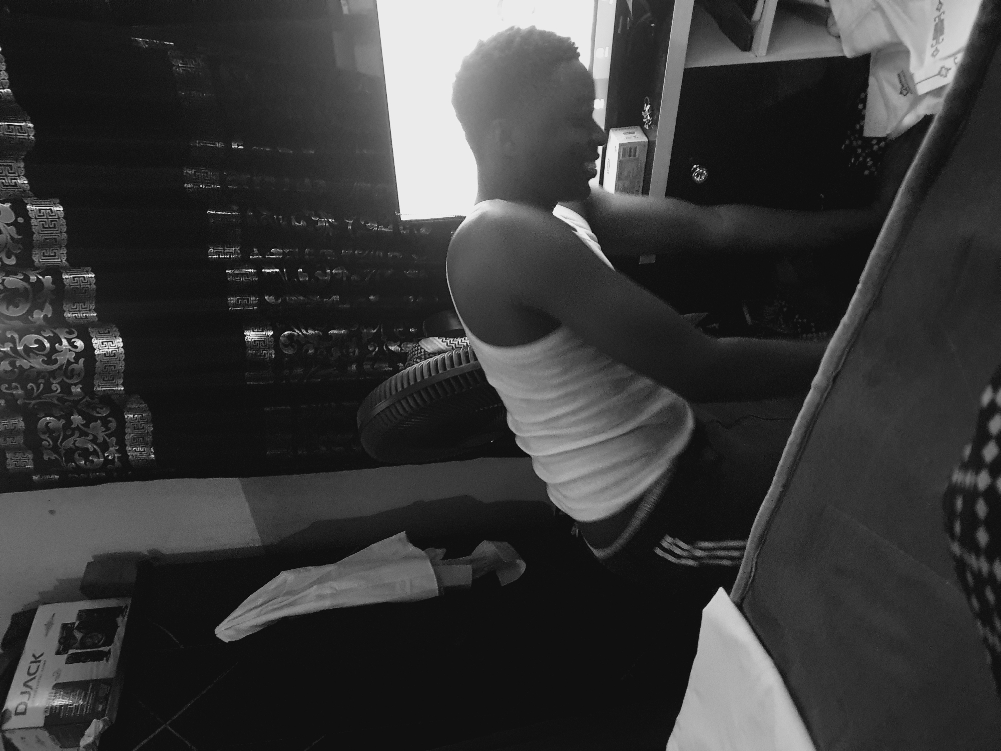 The image shows a person in a white tank top and black shorts sitting. The background includes patterned curtains, a window, a fan, and a box labeled "D.JACK." The location is Lokoja, Nigeria, based on information provided.