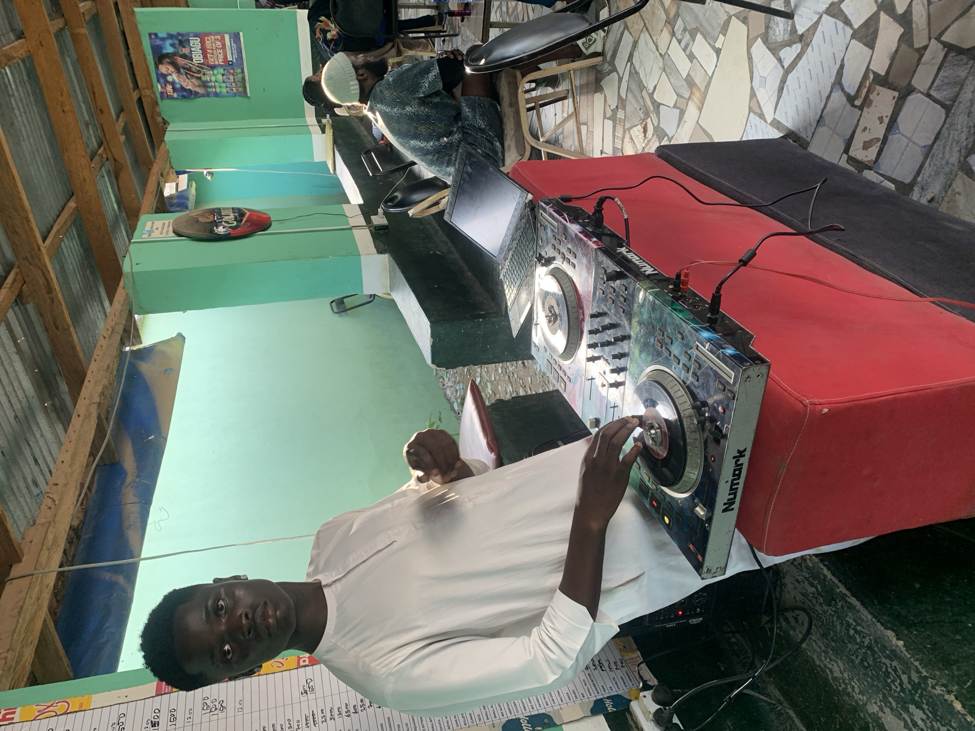 The image captures a daytime scene at a semi-open-air establishment in New Stadium, Nigeria, featuring a young male DJ positioned centrally. The DJ, with dark skin and short dark hair, wears a white, long-sleeved tunic and looks directly at the camera. His right hand rests on a jog wheel of a multi-colored Numark DJ controller, which is placed on a red upholstered stand. A black laptop with a dark screen is open behind the controller on the same stand.
To the DJ's left, a tall white board lists handwritten items and prices, including "MALTINA," "GULDER," "ICE," and other products. The background consists of light green painted concrete walls and pillars supporting a corrugated metal roof with visible wooden support beams and sections of blue tarp. A circular "Golden Guinea" beer sign is mounted on one pillar, and a "LIFE OBIAGU" poster advertising "Get 4 60CL Bottles for the price of 3" is affixed to another wall.
In the mid-ground, to the right of the DJ, a man with a beard and a light-colored beanie is seated at a small table, facing away from the camera. He wears a dark, patterned short-sleeved top. Other individuals are partially visible further in the background. The floor is paved with irregularly shaped tiles in various light and dark shades. Black metal chairs with padded seats are scattered around the tables. Power and audio cables are visible connected to the DJ equipment, trailing down the red stand to the floor.