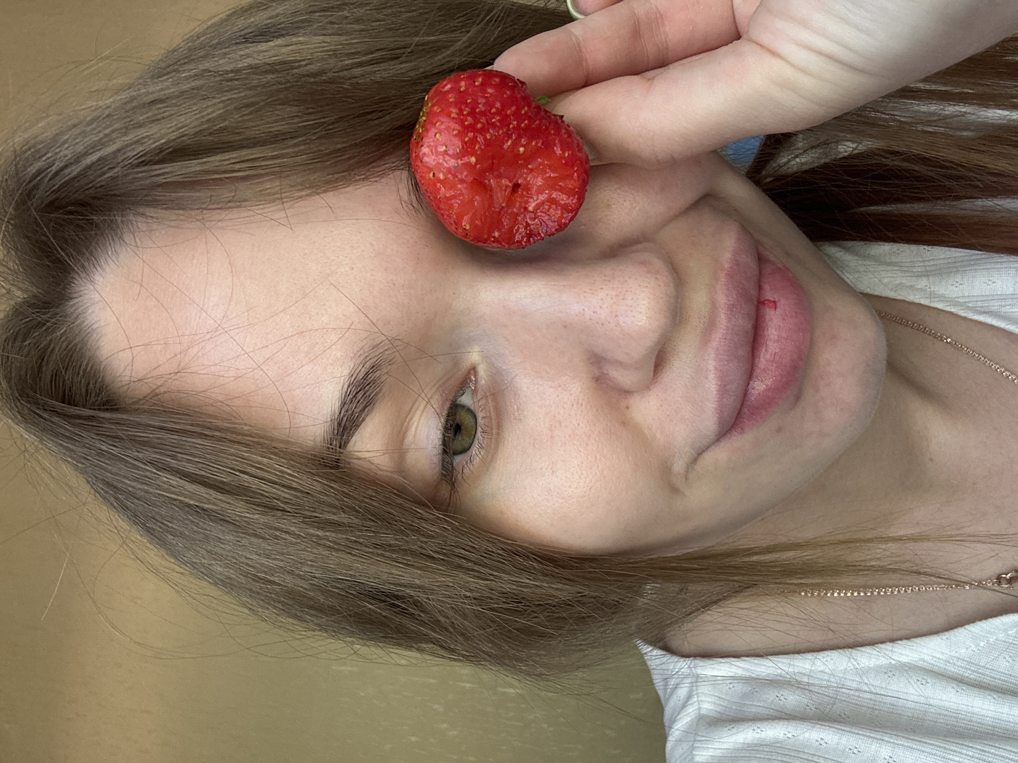 A close-up portrait depicts a female subject indoors, located in the city of Chaikovsky, Russia. The individual has long, light brown hair, some strands framing her face. Her visible left eye (viewer's right) is green, with dark eyeliner. Her right eye (viewer's left) is completely obscured by a single, red, ripe strawberry held in her right hand. She wears a white, ribbed top and a thin, gold-colored chain necklace. Her lips are full and slightly parted in a subtle smile or smirk, with a small red mark present on her lower lip. The background is an undifferentiated, light brown wall. The scene is illuminated by soft, even lighting.