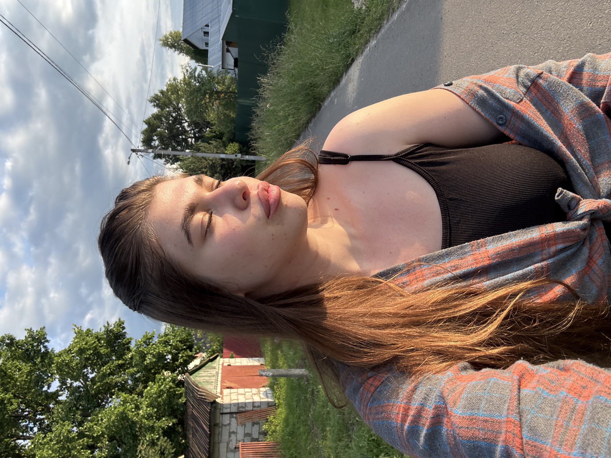 A young woman with long, brown hair is captured in a close-up selfie, her eyes closed as if basking in sunlight. She is wearing a black tank top and a plaid shirt, the sleeves of which are casually tied around her waist. Her skin is lightly tanned with freckles scattered across her cheeks. The scene appears to be outdoors in a rural setting, likely in Мещёры, Russia, given the context. In the background, a paved road runs alongside a grassy verge, with trees and simple houses visible. The sky is partly cloudy, suggesting it is daytime, possibly late afternoon due to the warm, golden light illuminating the scene. The overall mood is relaxed and peaceful, with the woman seemingly enjoying a moment of quiet reflection outdoors. No visible text is present in the image.
