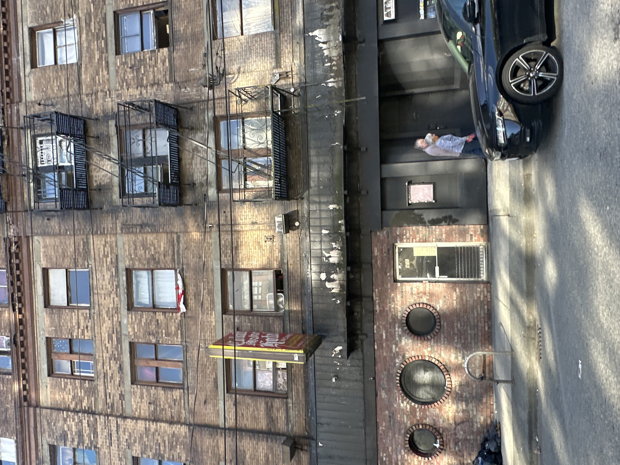 A brick building in an urban setting dominates the left side of the image. The building has multiple windows, some with decorative metal bars or fire escapes. A black car is parked near the entrance of the building, with a person standing near the car. The environment is urban, with a sidewalk and street visible on the right. Based on the architecture and signage, it might be Vancouver, Canada.