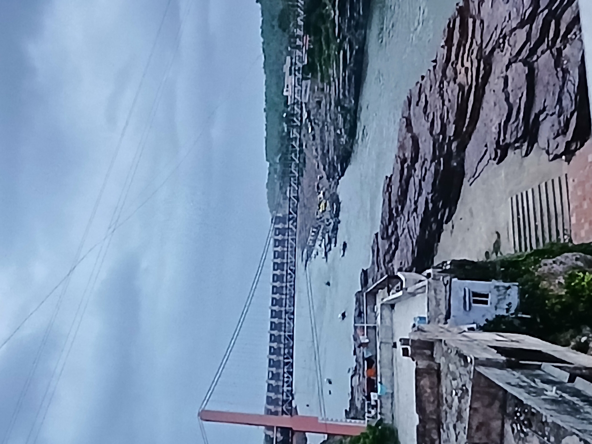 An aerial view of a bridge crossing a river in Rishikesh, India. The bridge is a large metal structure, with support towers on each side. The river is wide and calm, with green banks. There are buildings on the banks of the river, including a hotel and a temple. The sky is cloudy and overcast.