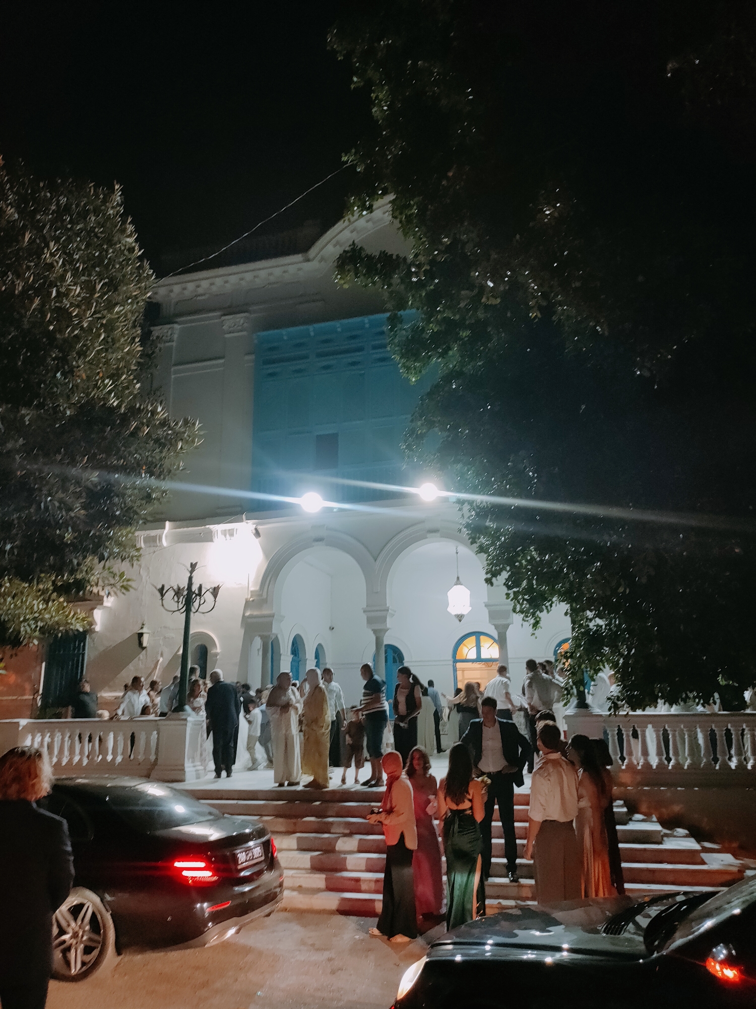 The image captures an evening scene outside a white building with arched entrances and windows, likely a public building or a historical site in Tunis, Tunisia. A group of people, dressed in formal attire including gowns and suits, are gathered on a set of wide stairs leading up to the building. Some are talking, while others appear to be arriving or departing. A black car is parked in the foreground on the right, and another black car is parked on the left, with its taillights illuminated. Trees with dark foliage are visible on the left and right sides of the frame, obscuring parts of the building and the sky. The lighting suggests it is nighttime, with artificial lights illuminating the area, creating a festive or event-like atmosphere. The architecture features classical elements, with decorative railings and columns. A traditional-style street lamp is visible on the left side of the stairs.