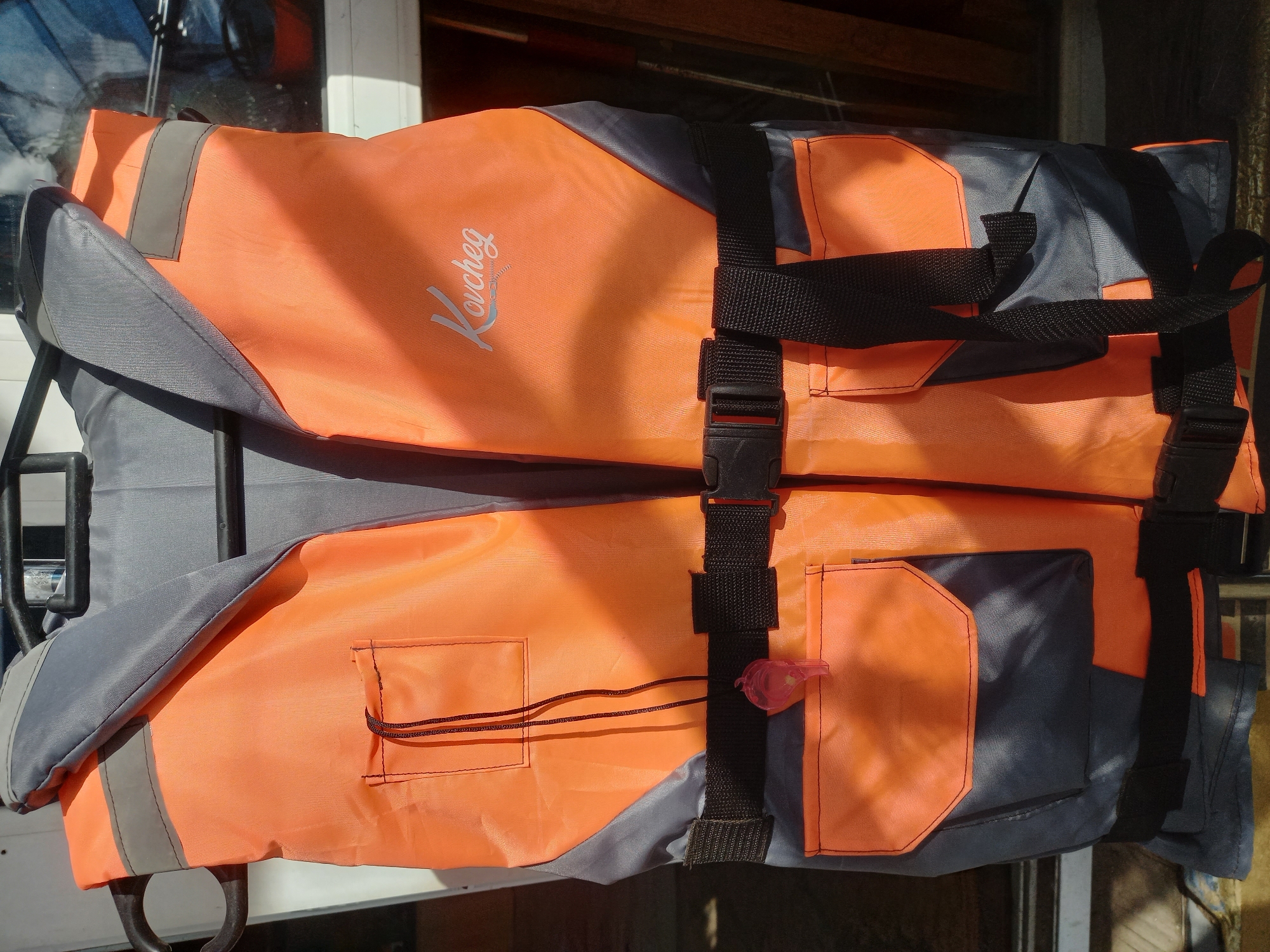 The image shows a folded orange and gray life jacket with black straps and buckles. The jacket has the word "Kovcheg" printed on the orange fabric. A small pocket is visible on one side, with a pink whistle attached to a string. The life jacket appears to be placed on a surface, possibly a boat. The location is unknown.