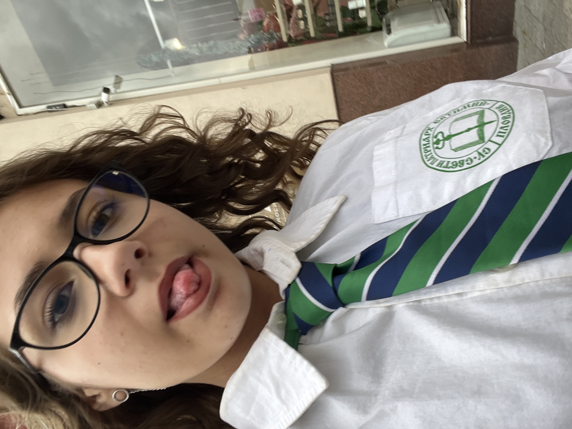 A young woman is captured in a close-up, upward-angle selfie, likely taken in Plovdiv, Bulgaria. She has dark, curly hair and wears black-framed eyeglasses. Her expression is playful, with her tongue slightly out. She is dressed in a white collared shirt and a striped tie featuring green, navy blue, and white diagonal lines, suggesting a school uniform. A small silver hoop earring is visible on her left ear.
A distinct circular green logo is prominent on the right shoulder area of her shirt. The Cyrillic text within the logo reads "ГУ-СВ.ПАТРИАРХ ЕВТИМИЙ - ПЛОВДИВ", which translates to "High School St. Patriarch Euthymius - Plovdiv". This text provides a specific location and institutional context for the image.
The background features reflections in what appears to be a large window or glass panel, indicating an outdoor or semi-outdoor setting. These reflections show an overcast or cloudy daytime sky and some indistinct indoor elements, possibly a counter or a cash register. The overall impression is a casual moment captured in an urban environment, likely near the high school identified by the uniform and logo.