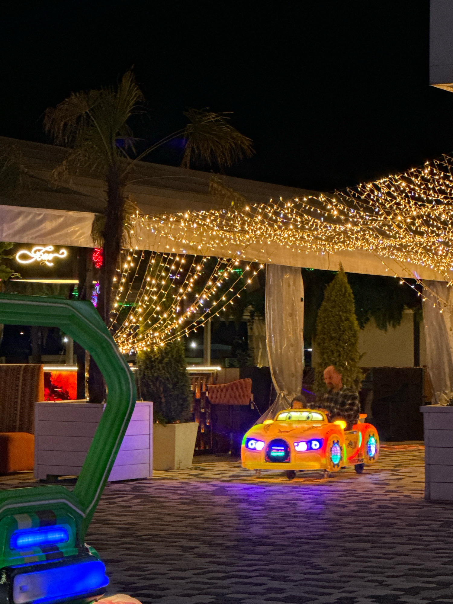 The image depicts a nighttime scene at a resort or entertainment venue in Lermontovo, Russia. A man is seated in a brightly colored, illuminated toy car, likely a ride for children, which is parked on a patterned outdoor floor. Overhead, strings of warm yellow lights are strung across a canopy, illuminating the area. Palm trees are visible in the background, suggesting a warm climate. The overall atmosphere is festive and recreational, with decorative lighting and amusement rides.