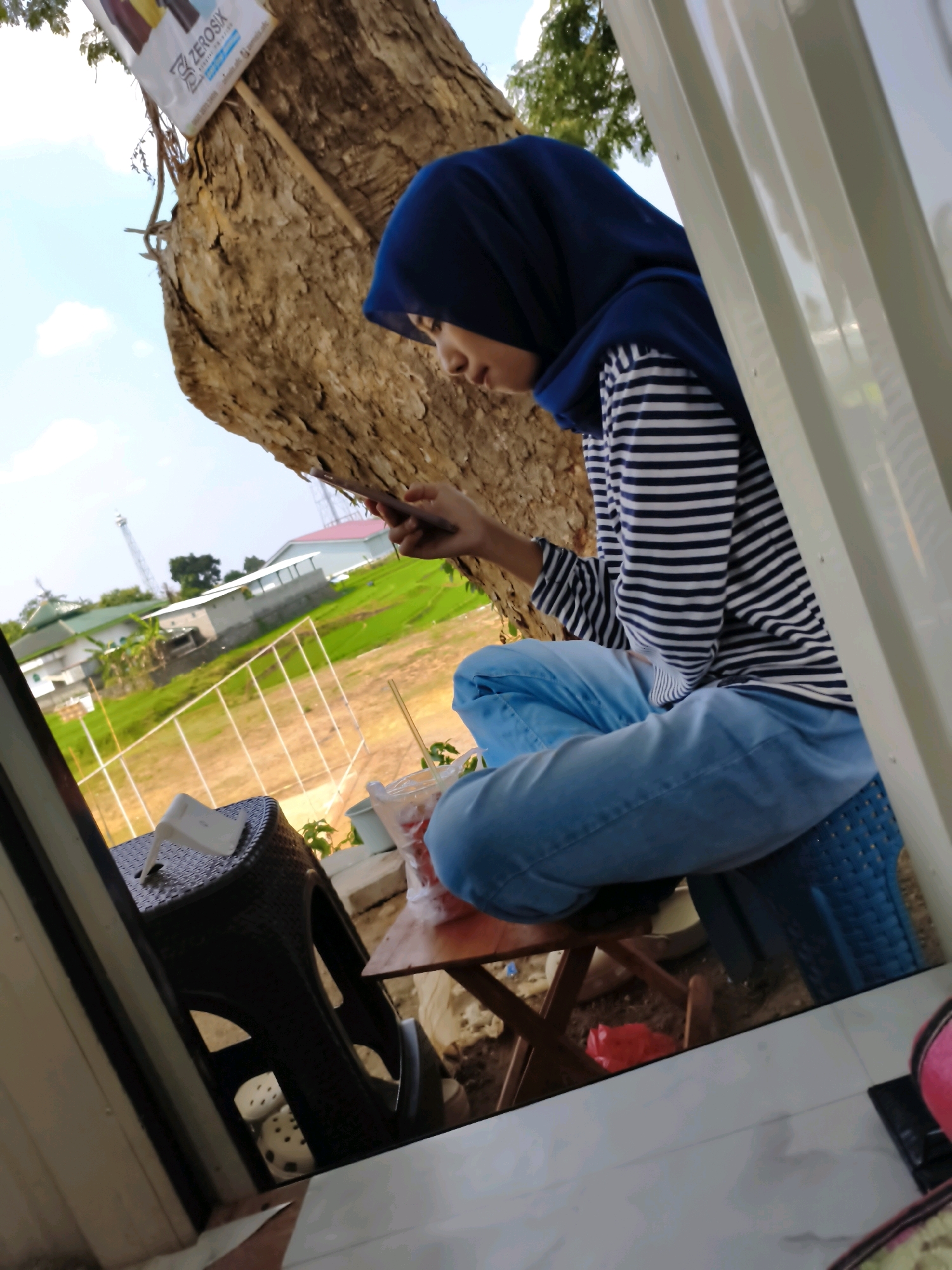 A young woman wearing a navy blue hijab and a horizontally striped long-sleeved shirt sits cross-legged on a blue plastic stool. She is looking down at a mobile phone in her hands. She is wearing light blue jeans. The woman is positioned next to a large tree with rough bark, under which is a small wooden table. On the table are a few items, including a white tissue box, and a plastic cup with a straw and some red liquid. In the background, a rural landscape is visible, with green rice paddies, some buildings, and a communication tower. The scene appears to be set in a cafe or outdoor seating area. The location is Puhpelem, Indonesia.