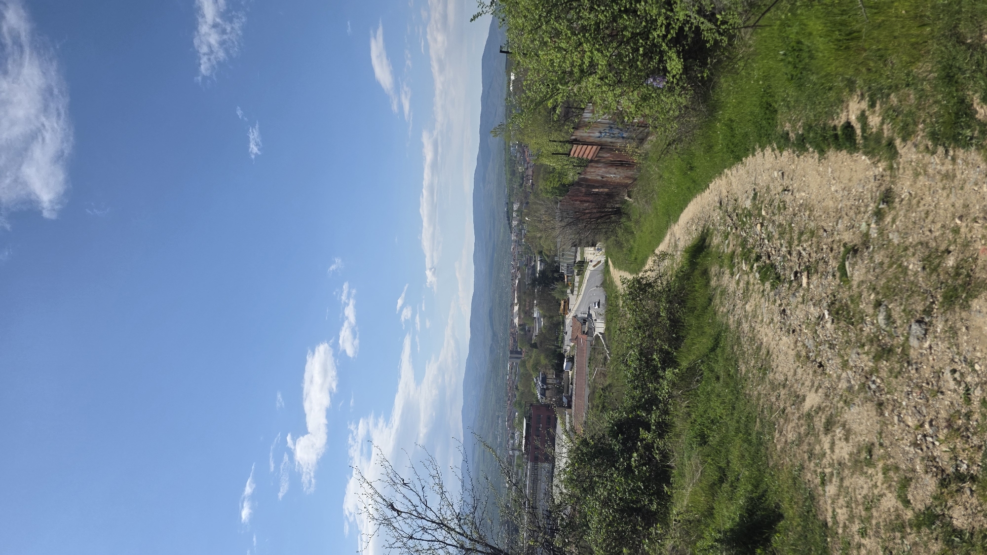 The image shows a view from a hill overlooking the city of Blagoevgrad, Bulgaria. A dirt path runs along the right side of the frame, lined with green grass and vegetation. A rusty fence is visible along the path. In the distance, the cityscape of Blagoevgrad is visible, with buildings, streets, and cars. Mountains form the backdrop of the city. The sky is blue with scattered white clouds. Bare tree branches are visible in the foreground. The overall perspective is elevated.