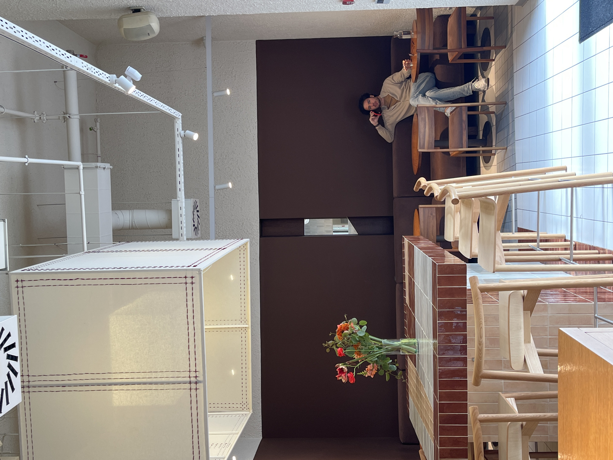 A man is lying on a dark brown couch next to a wooden table and sitting area. The man is holding a phone up to his ear. In the lower left corner of the image is a white storage cube. There is a bouquet of flowers on the table. The location appears to be Belgrade, Serbia.