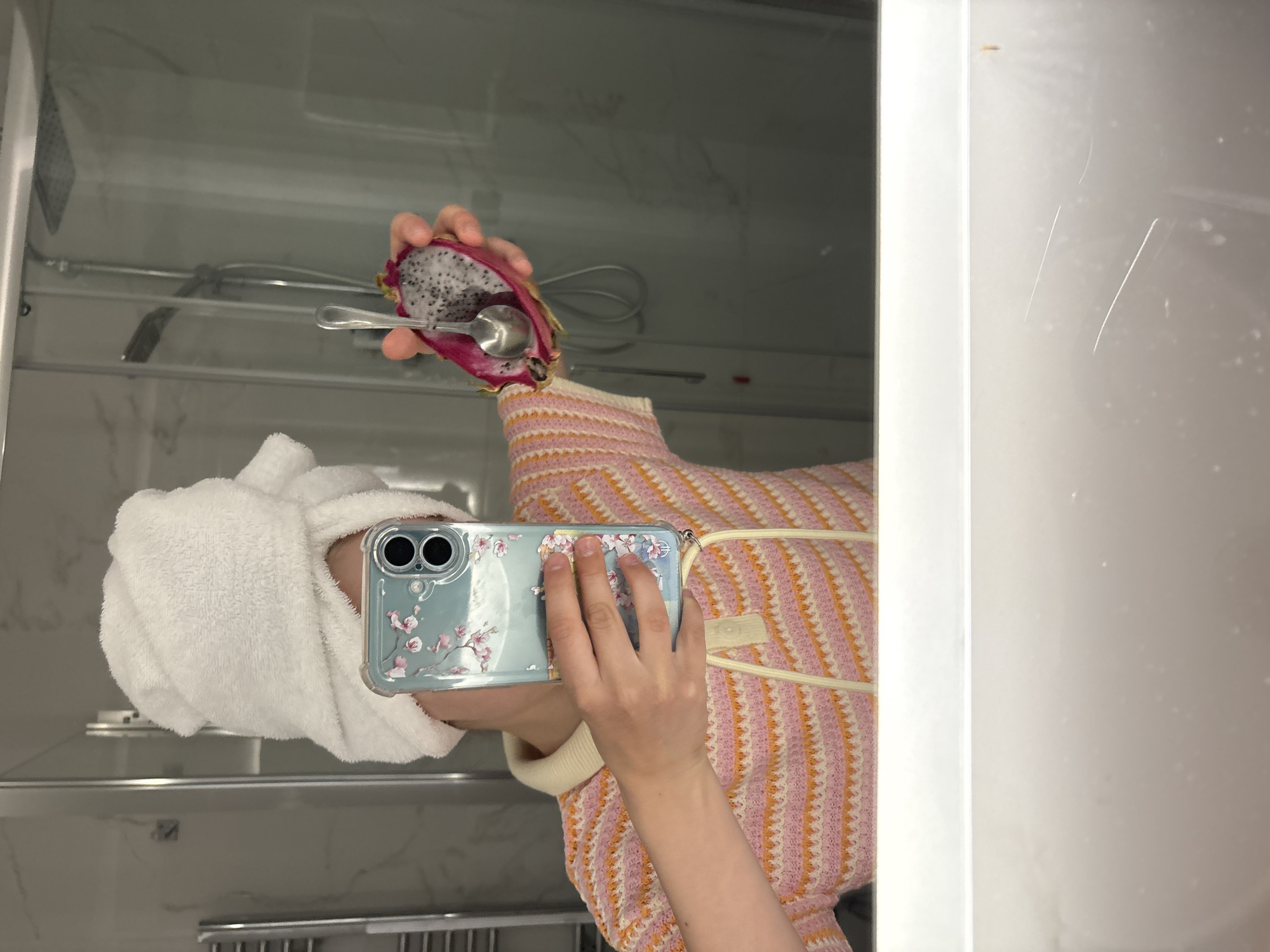 A person with a towel wrapped around their head is reflected in a mirror, holding a phone in their hands and taking a picture. The phone has a transparent case decorated with cherry blossoms. The person is wearing a pink and orange striped sweater. In the mirror, the person is also holding a halved dragon fruit with a spoon in it. The background includes a shower stall with a marble-like wall. The setting appears to be a bathroom. There's no indication of the location being Montecatini Terme, Italy, in the image.