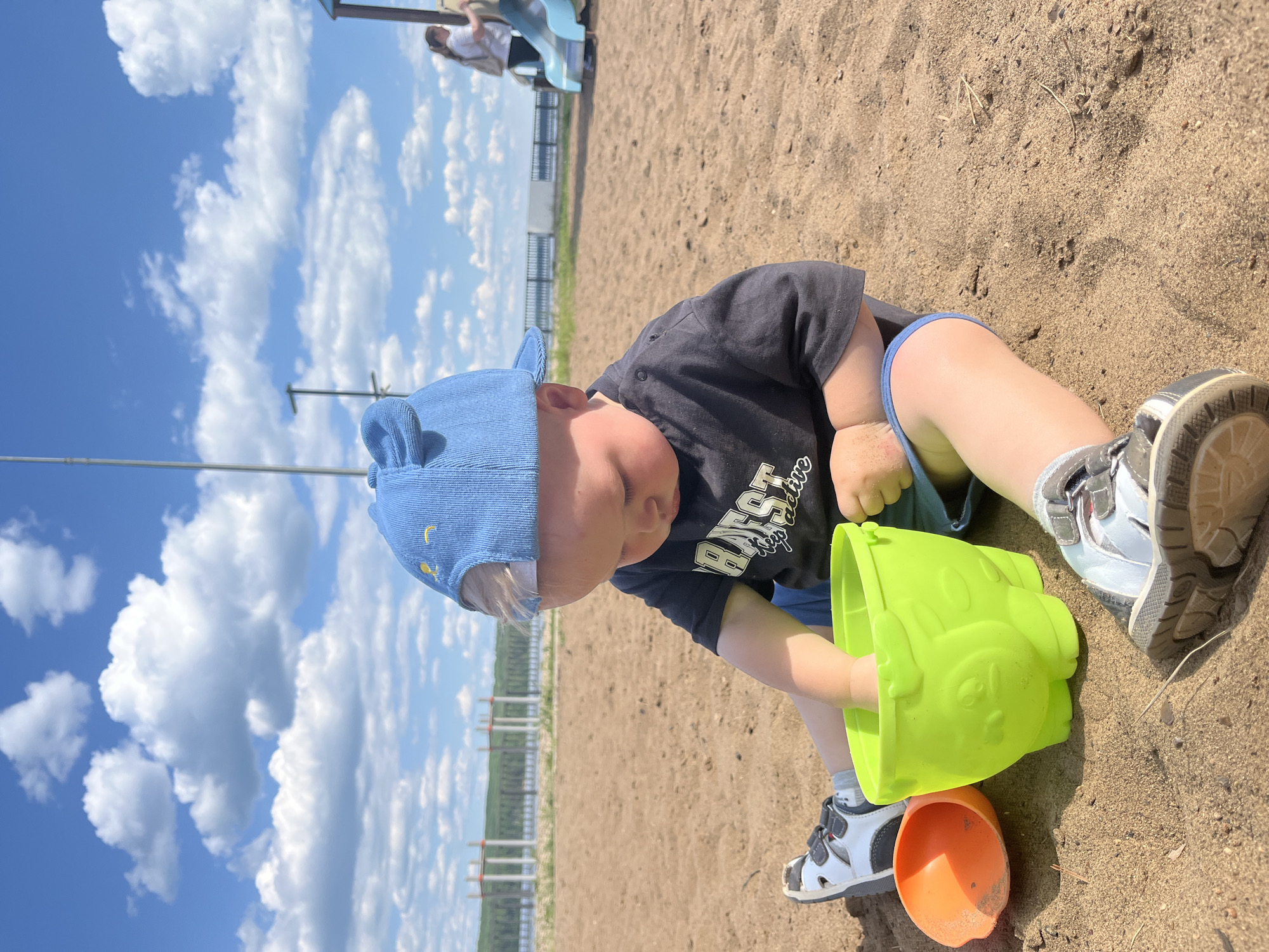 A young child wearing a blue baseball cap with bear ears and a black t-shirt sits in the sand at a playground. The child is holding a green plastic bucket in their hands, digging in the sand. An orange plastic shovel is to the left of the bucket. The child is wearing white and grey sandals. In the background, a playground slide and a woman are visible. The sky is blue with white clouds. The scene is set outdoors, likely at a park or beach. The location is described as being in the city of Chaikovsky, Russia.