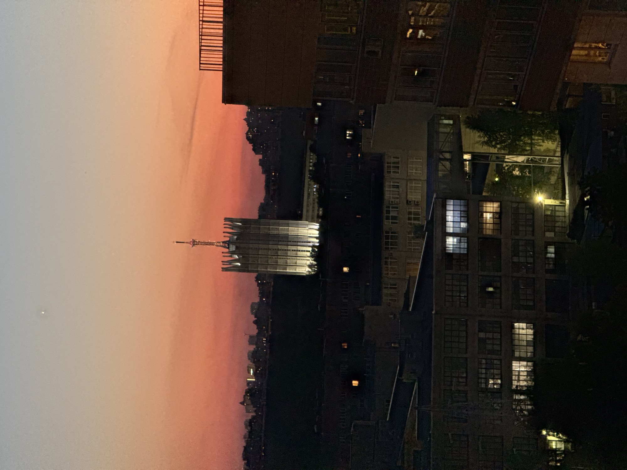 The image displays an urban cityscape at dusk or early night, captured from an elevated perspective. The sky occupies the upper portion of the frame, showcasing a striking gradient of colors: a pale, light greyish-blue fades into soft oranges and vibrant pinks closer to the horizon line. A small, bright point of light, likely a star or planet, is visible in the lighter part of the sky.
A prominent, tall building with an ornate, illuminated spire or antenna stands out against the colorful sky in the midground. Its intricate upper architecture and light-colored facade reflect the artificial light, making it a focal point. Surrounding this landmark, and extending into the foreground, are numerous dark, multi-story buildings, largely silhouetted against the ambient light. Many of their windows are illuminated, casting warm glows and suggesting ongoing activity within.
In the far distance, a sprawling city skyline forms a continuous dark silhouette along the horizon. Dark tree foliage partially obscures the far right side of the image. A dark, slatted structure, possibly a balcony railing, is visible in the top right corner, indicating the vantage point. The scene depicts a tranquil urban evening, with no explicit human activity visible. There is no visible text in the image.